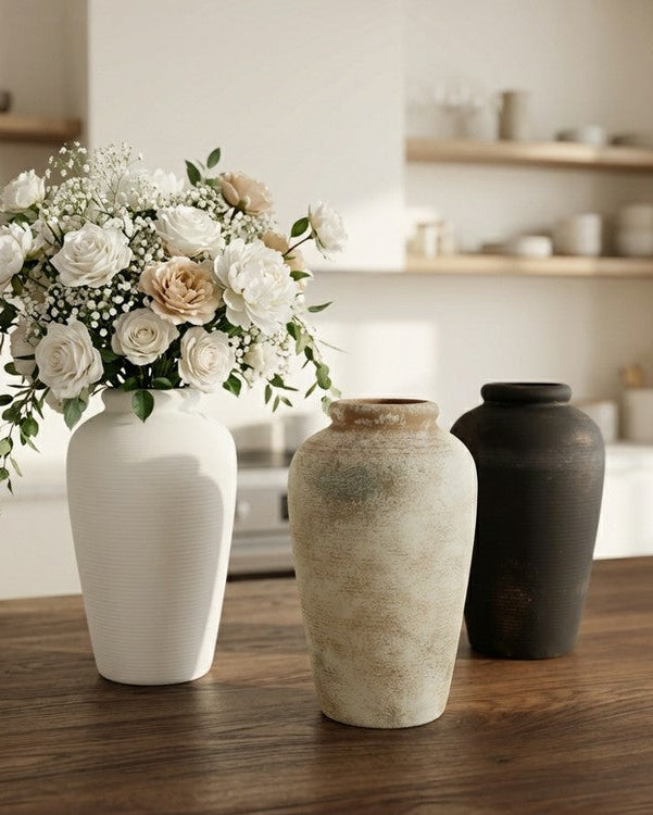 three ceramic vases in rust, white and black with flowers in one pot. on a wooden kitchen counter.