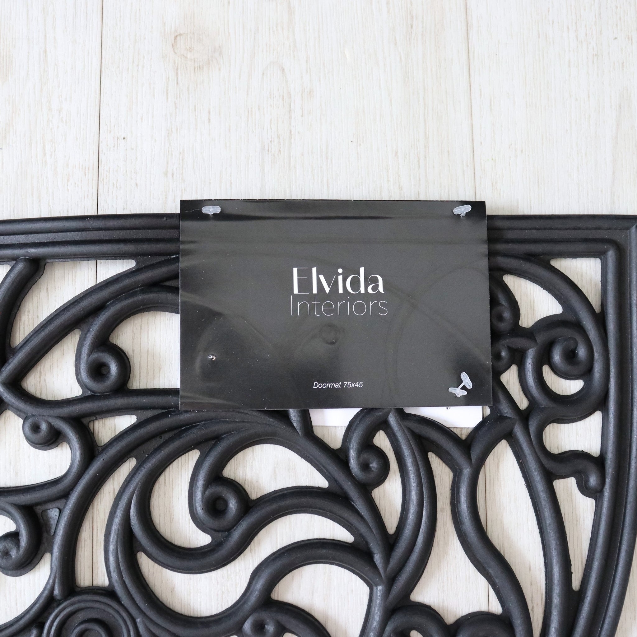 Decorative black metal doormat with 'Elvida Interiors' label on a light wooden floor.