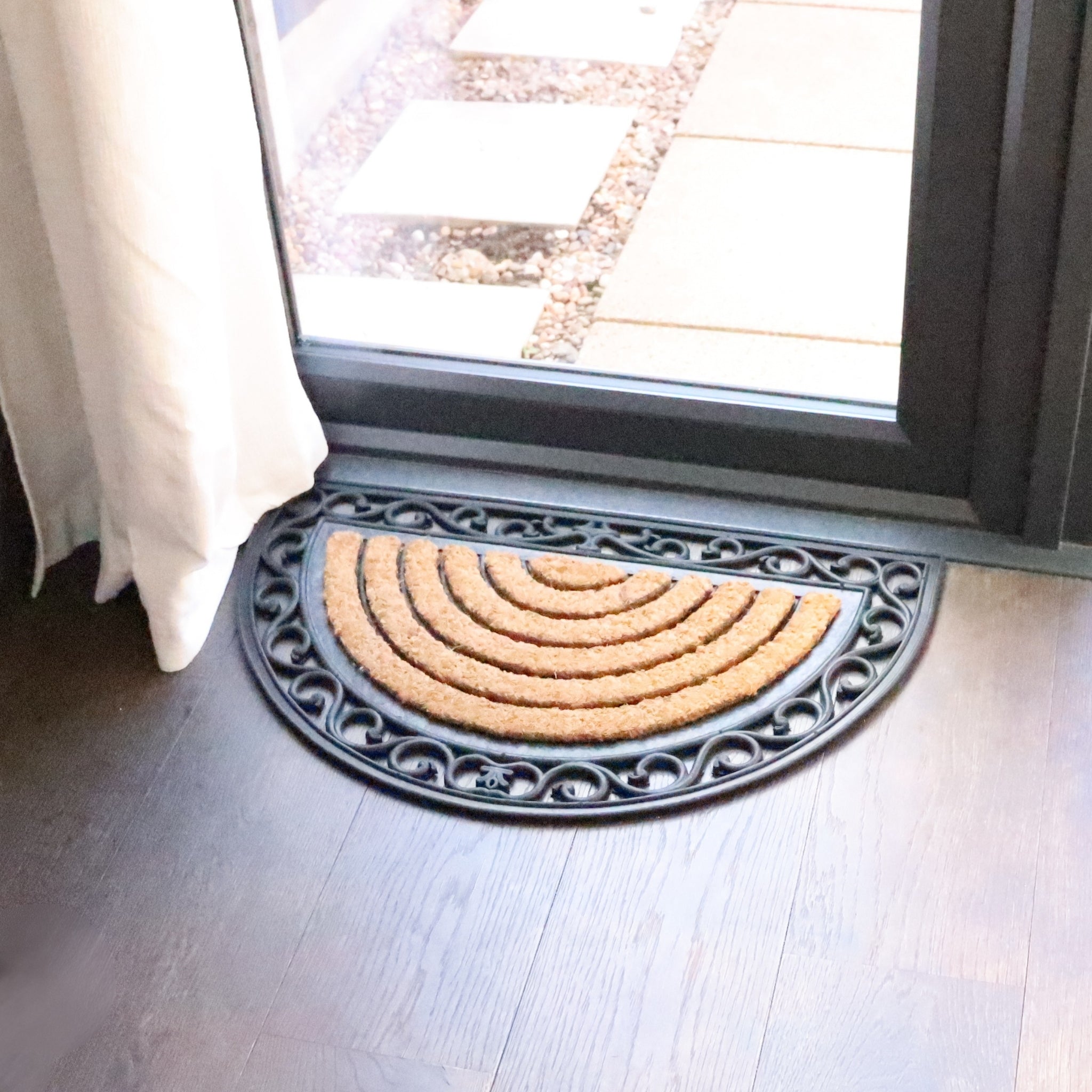 Half-circle doormat with decorative border on a wooden floor next to a glass door.