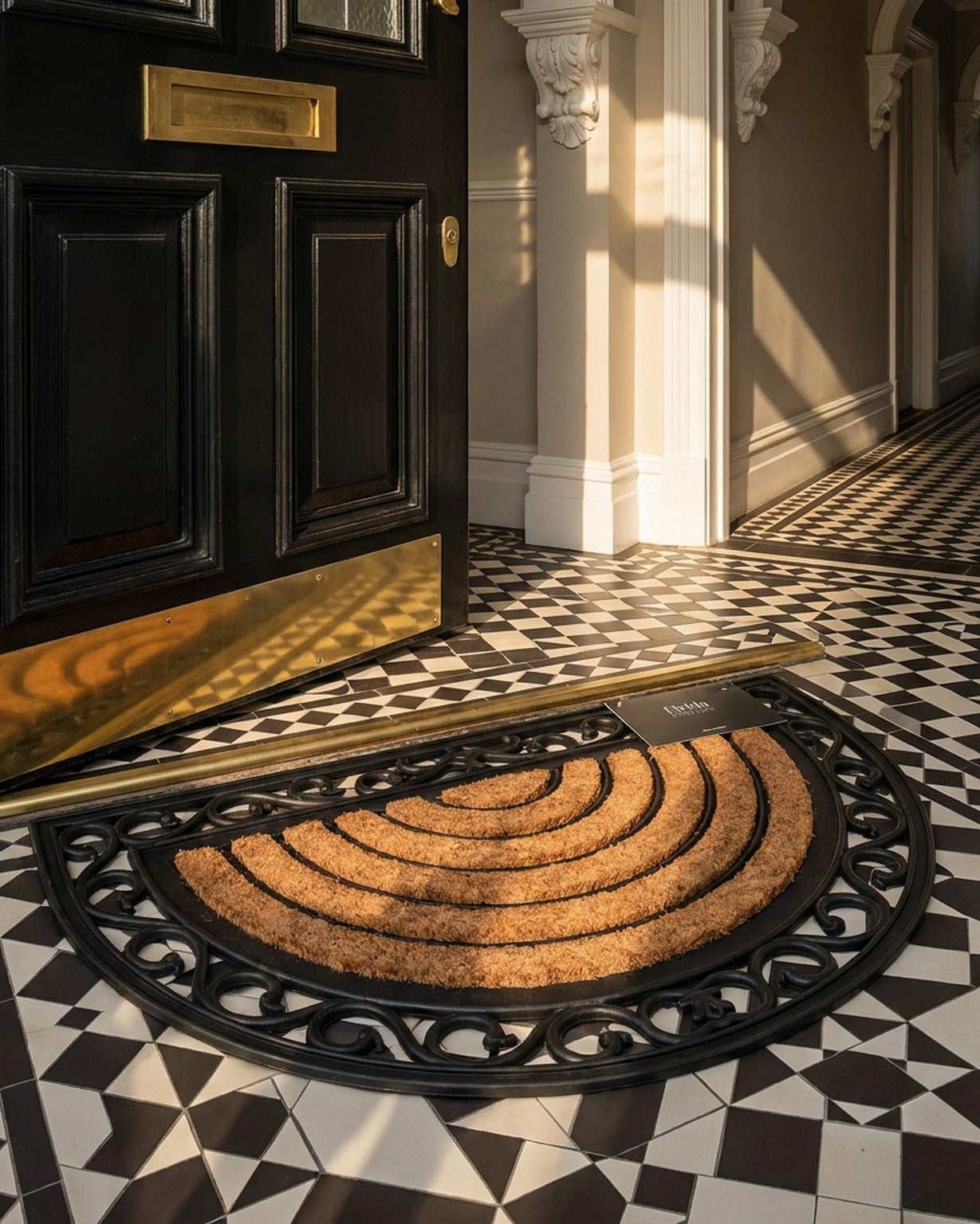 Doormat with geometric pattern in front of a black door with gold hardware on a black and white tiled floor.