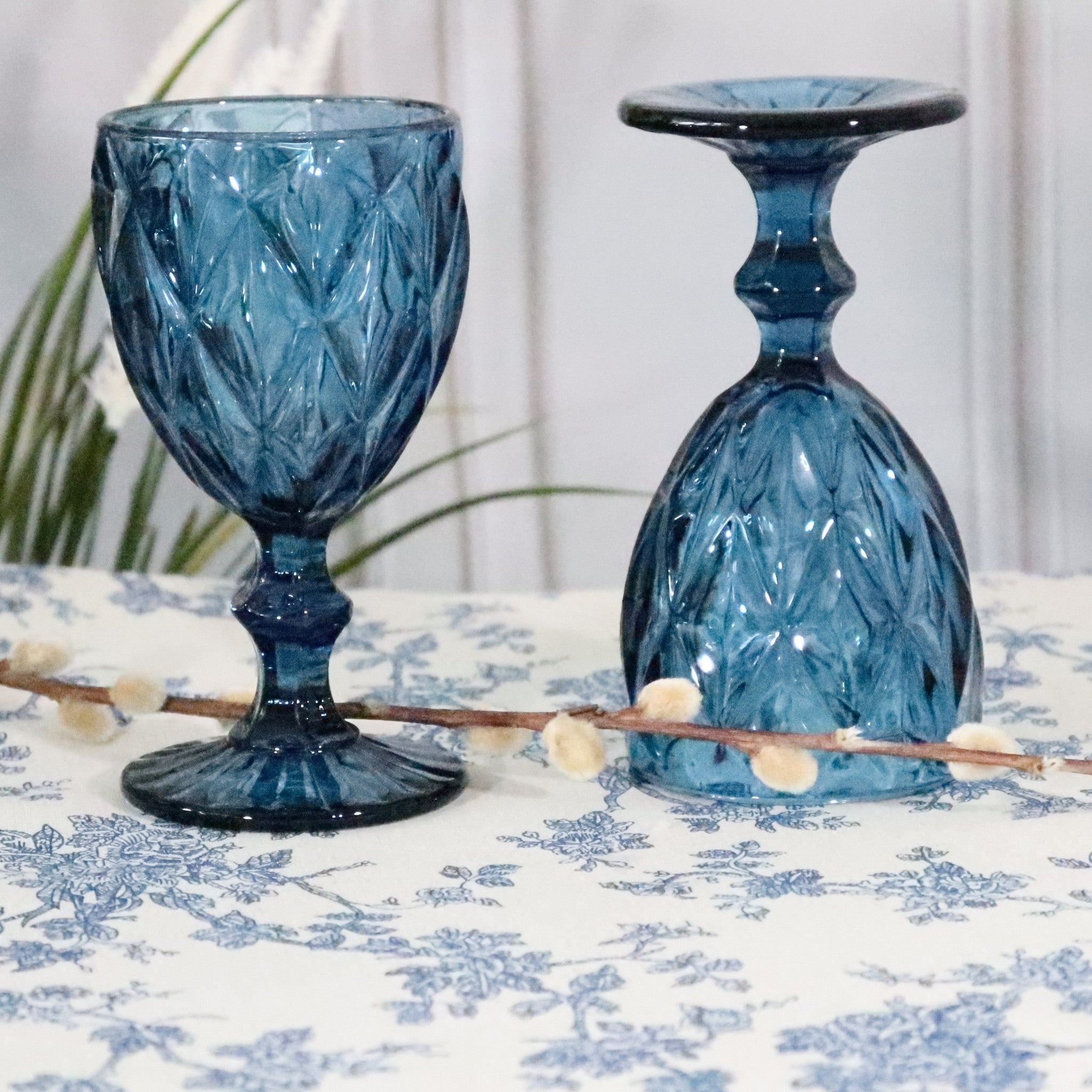 Two blue glass goblets on a floral tablecloth with a white curtain background