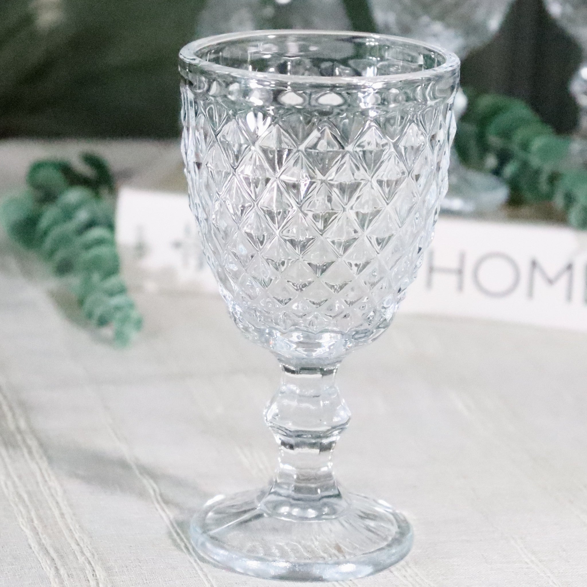 Clear glass goblet with diamond pattern on a light surface with greenery in the background