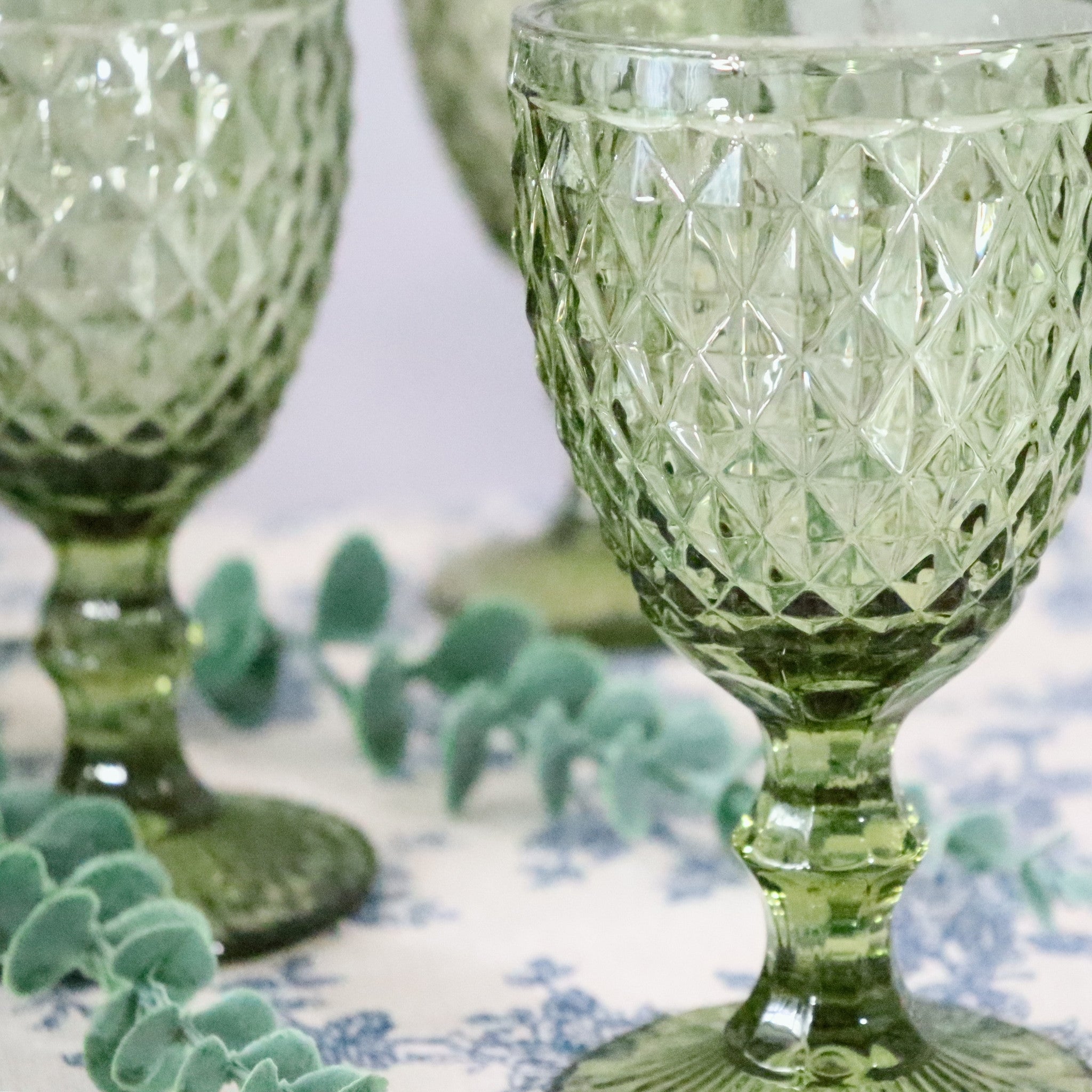 Green textured glass goblets on a patterned tablecloth with greenery.