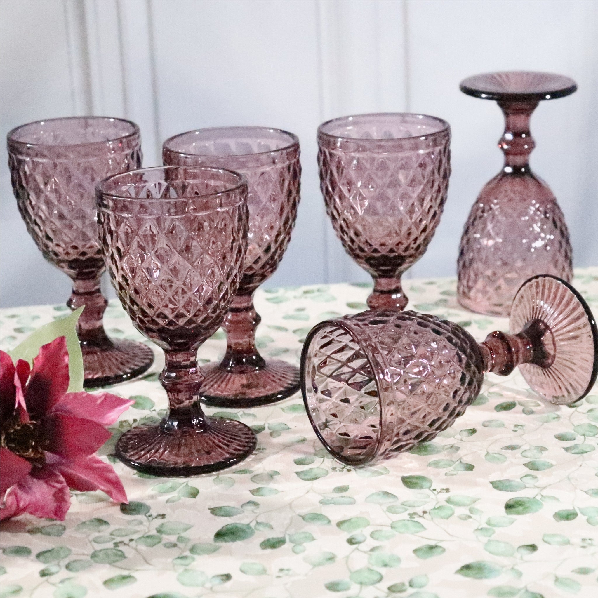 Set of purple glass goblets on a patterned surface with a floral arrangement.