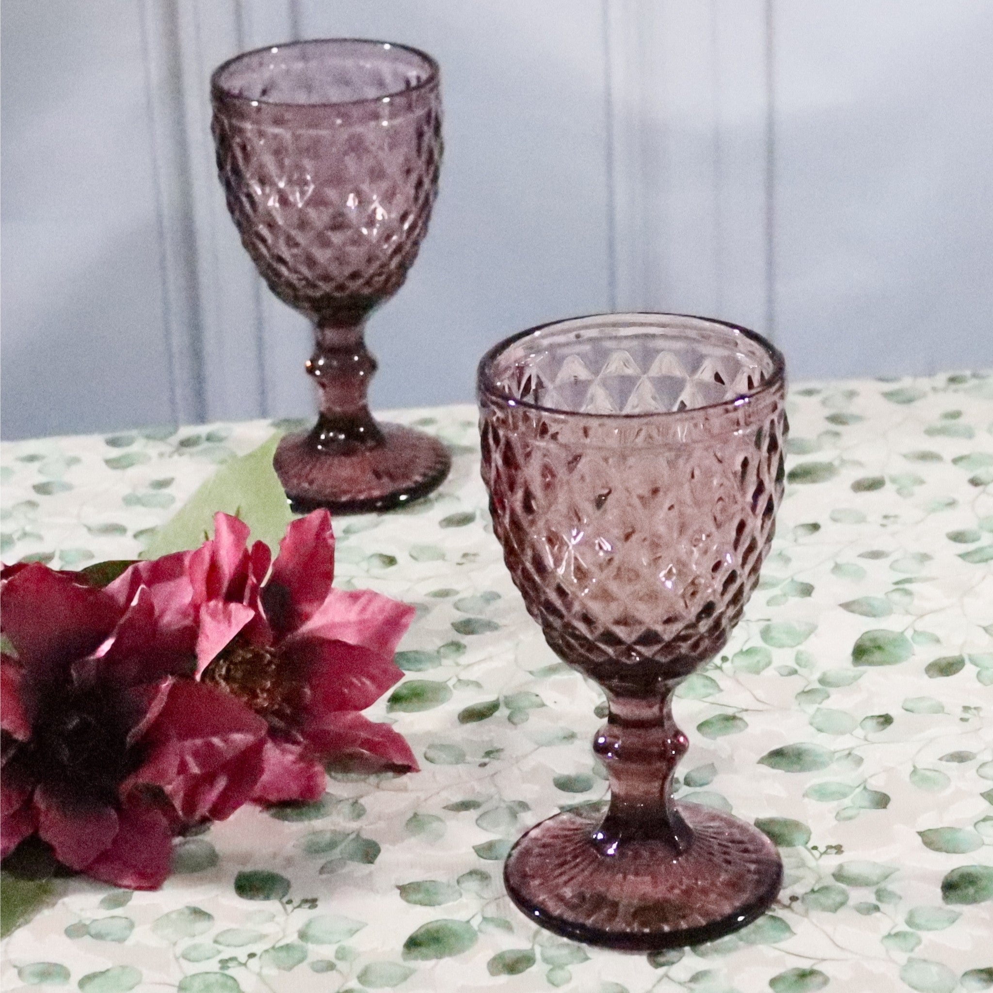 Two purple textured glass goblets on a table with a floral arrangement.