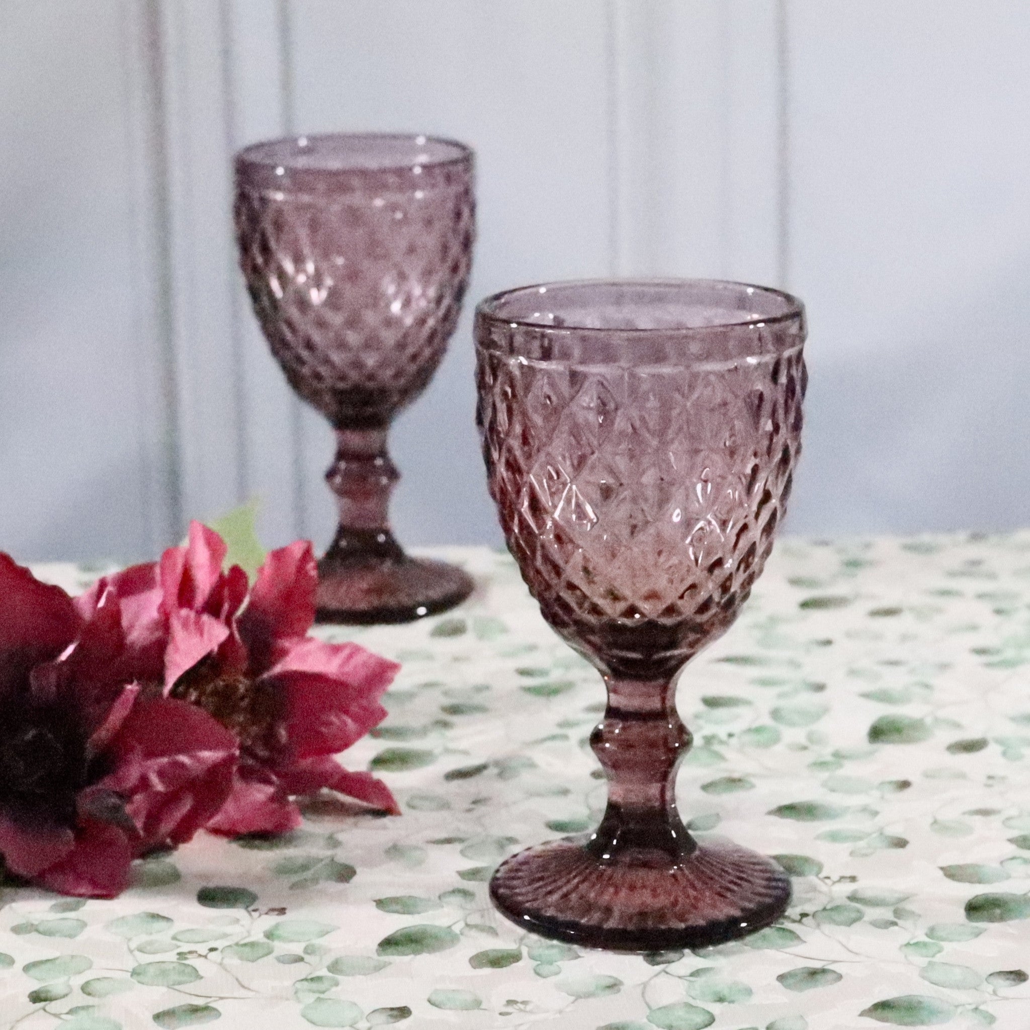 Two purple textured glass goblets on a patterned surface with flowers.