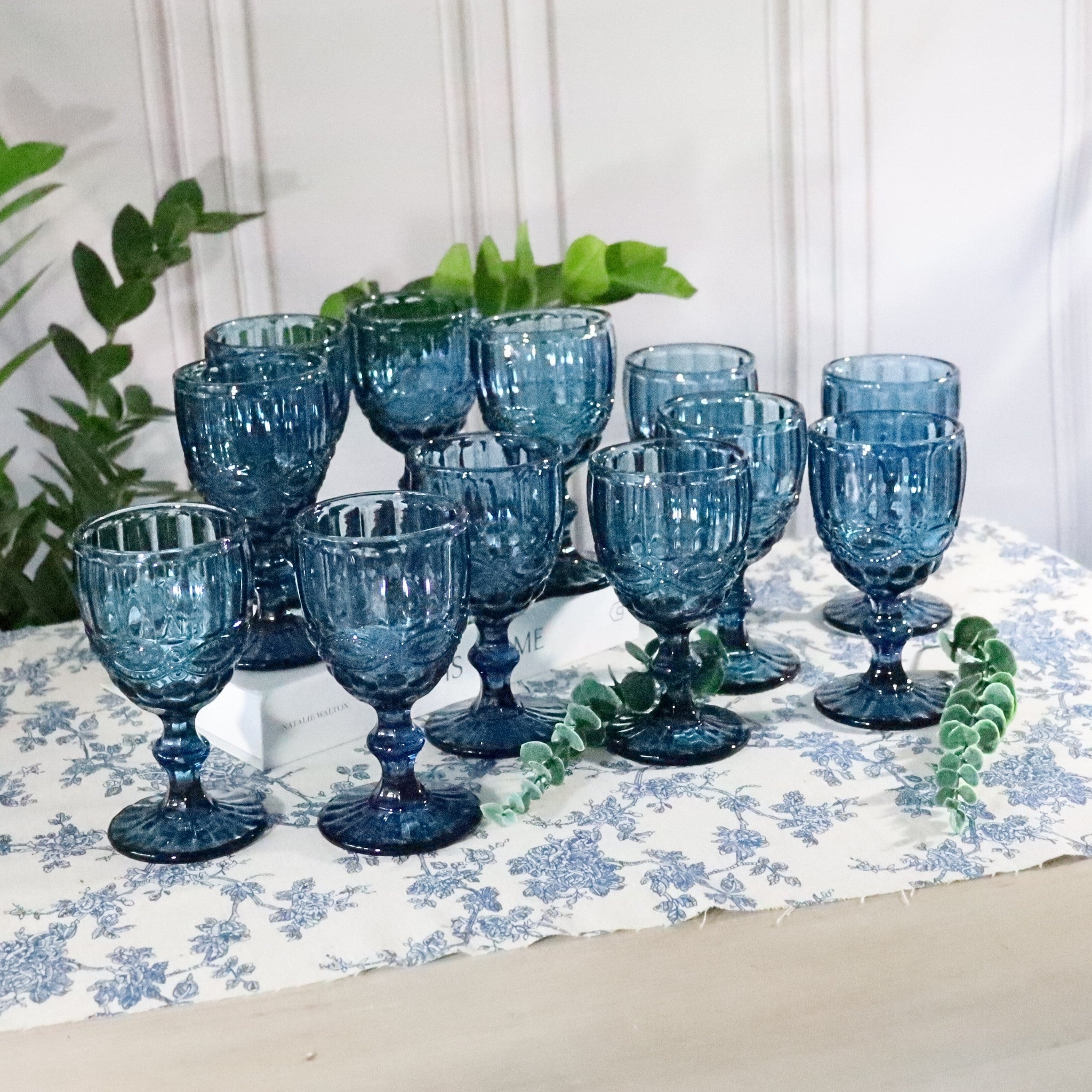 Set of blue glass goblets on a floral tablecloth with plants in the background