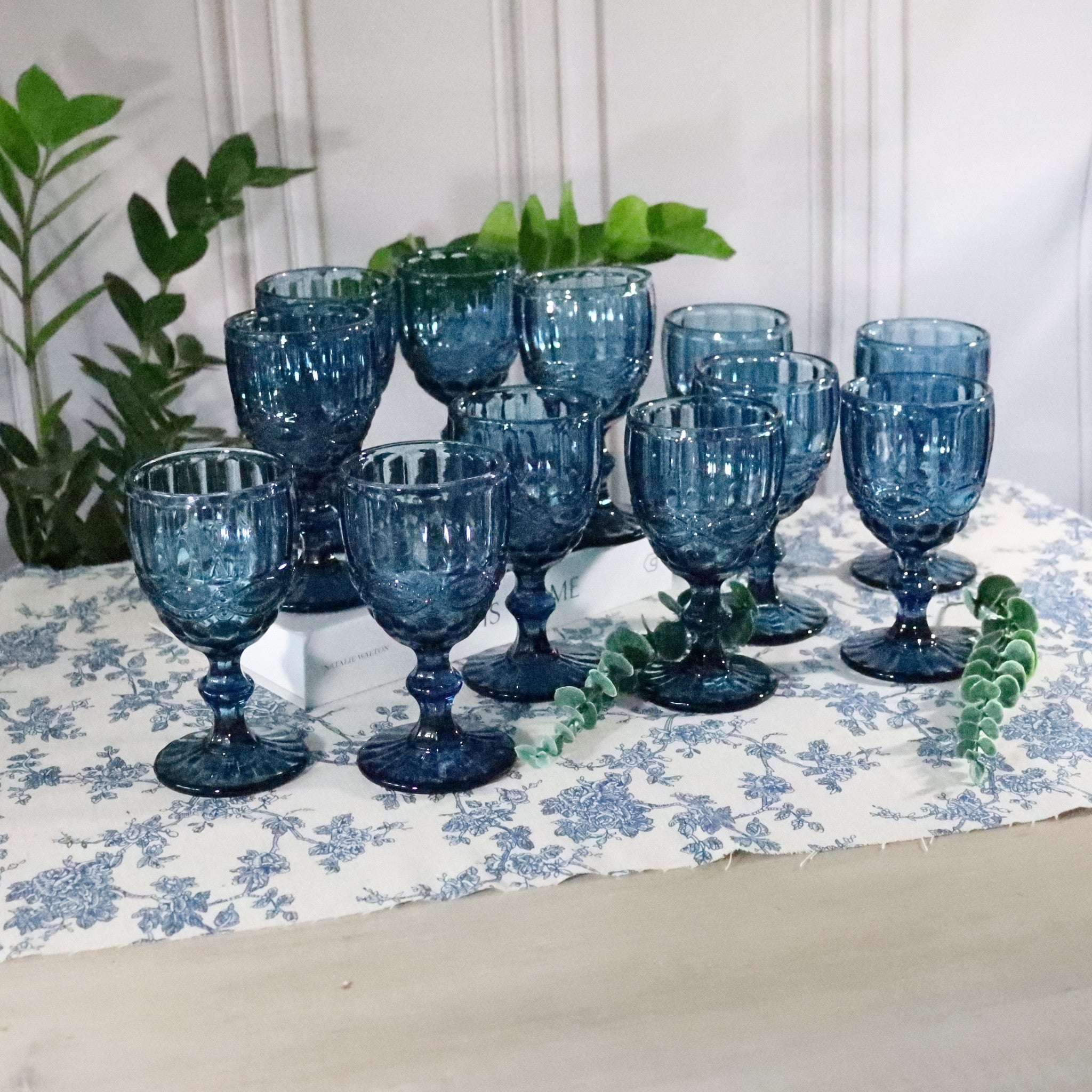 Set of blue glass goblets on a floral tablecloth with plants in the background