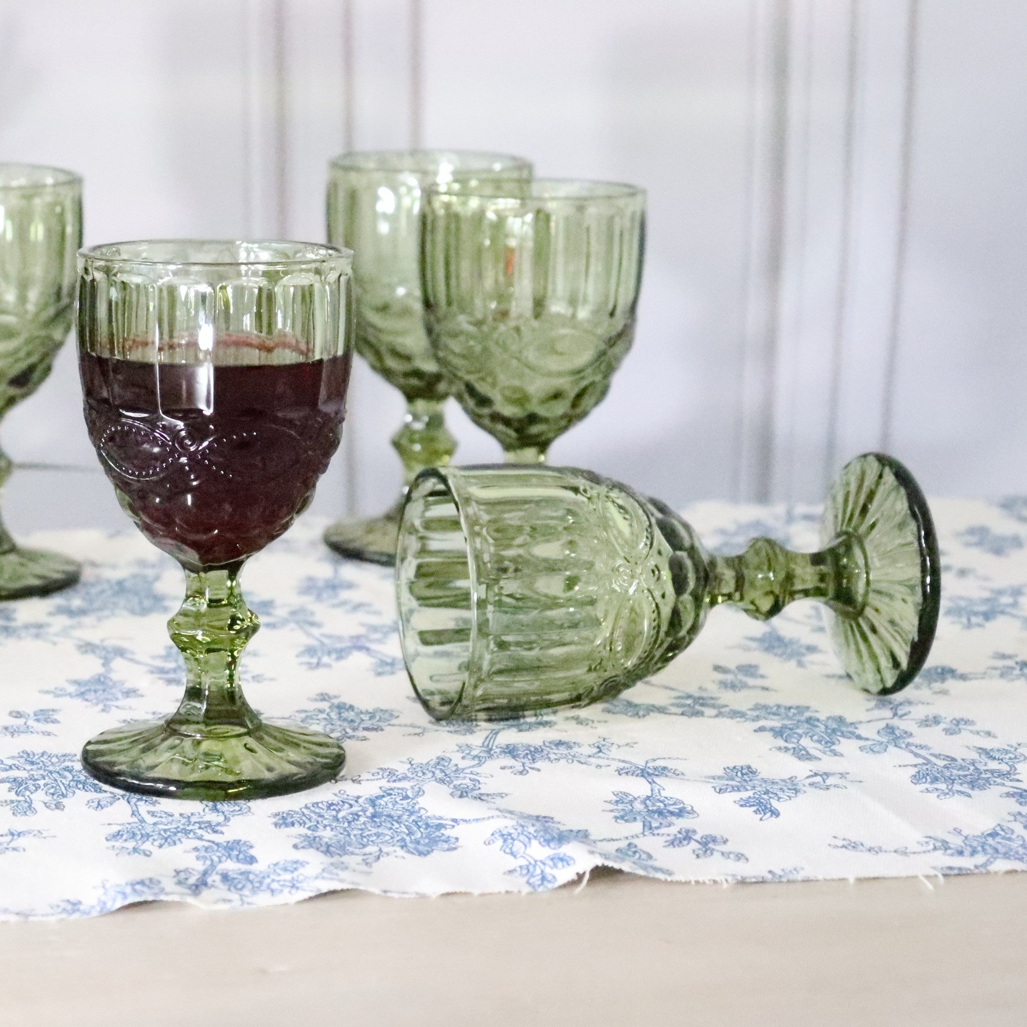 Green glass goblets on a floral tablecloth with one containing a dark liquid.