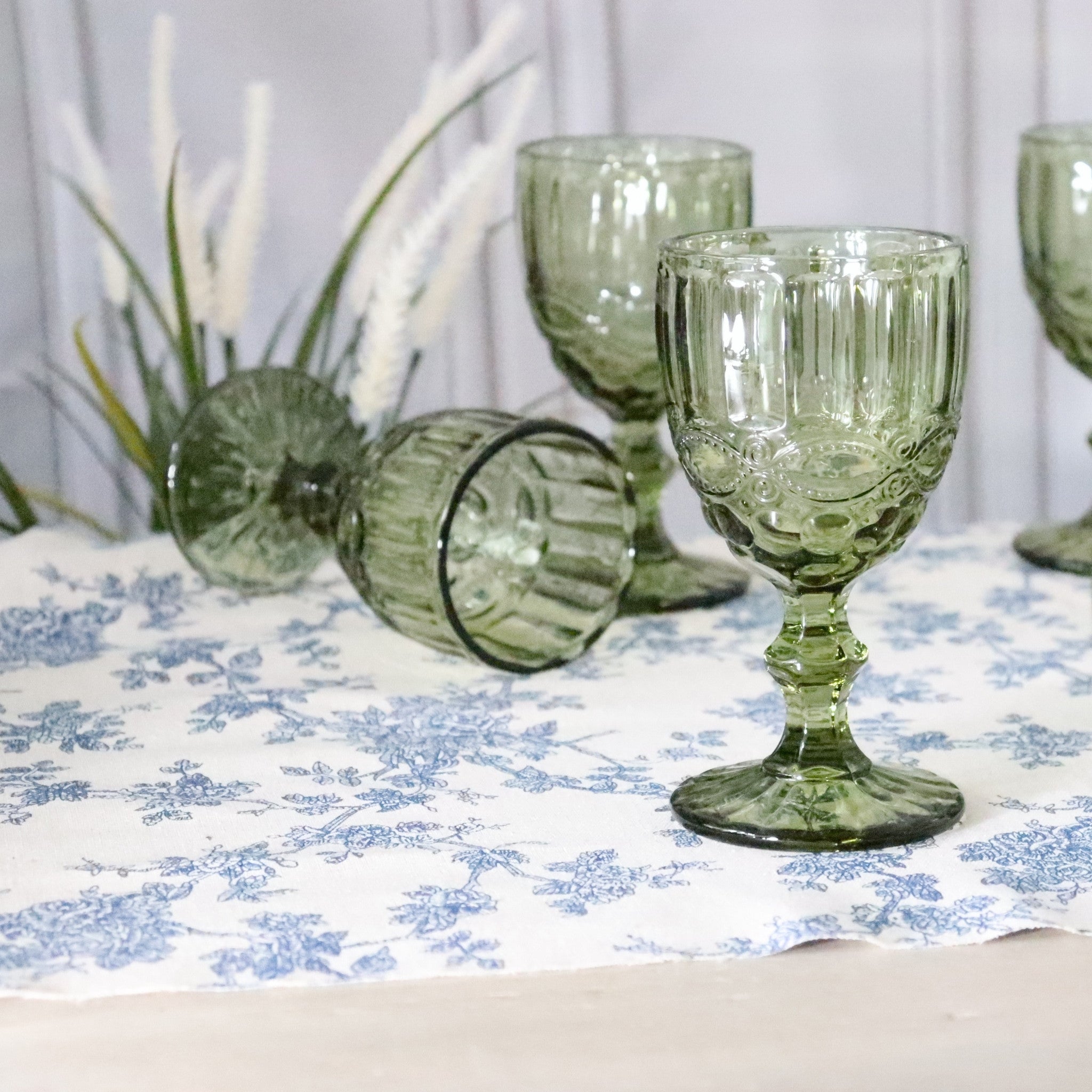 Green glass goblets on a floral tablecloth with a white curtain background