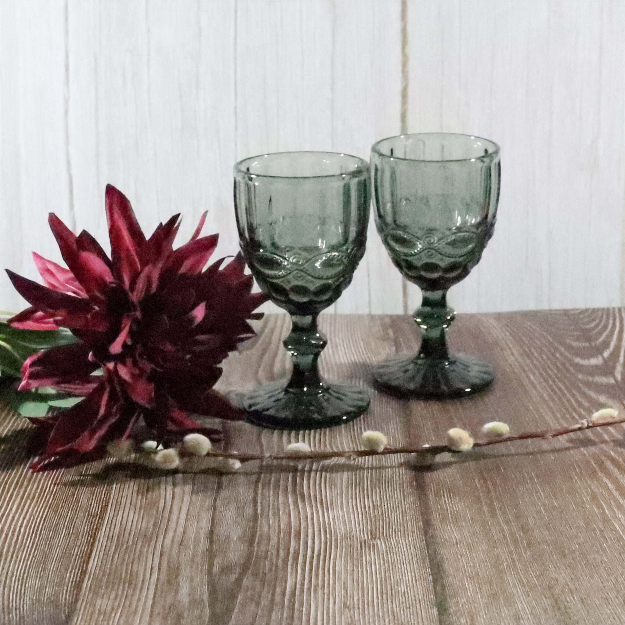 Two grey glass goblets on a wooden table with a decorative flower and branch.