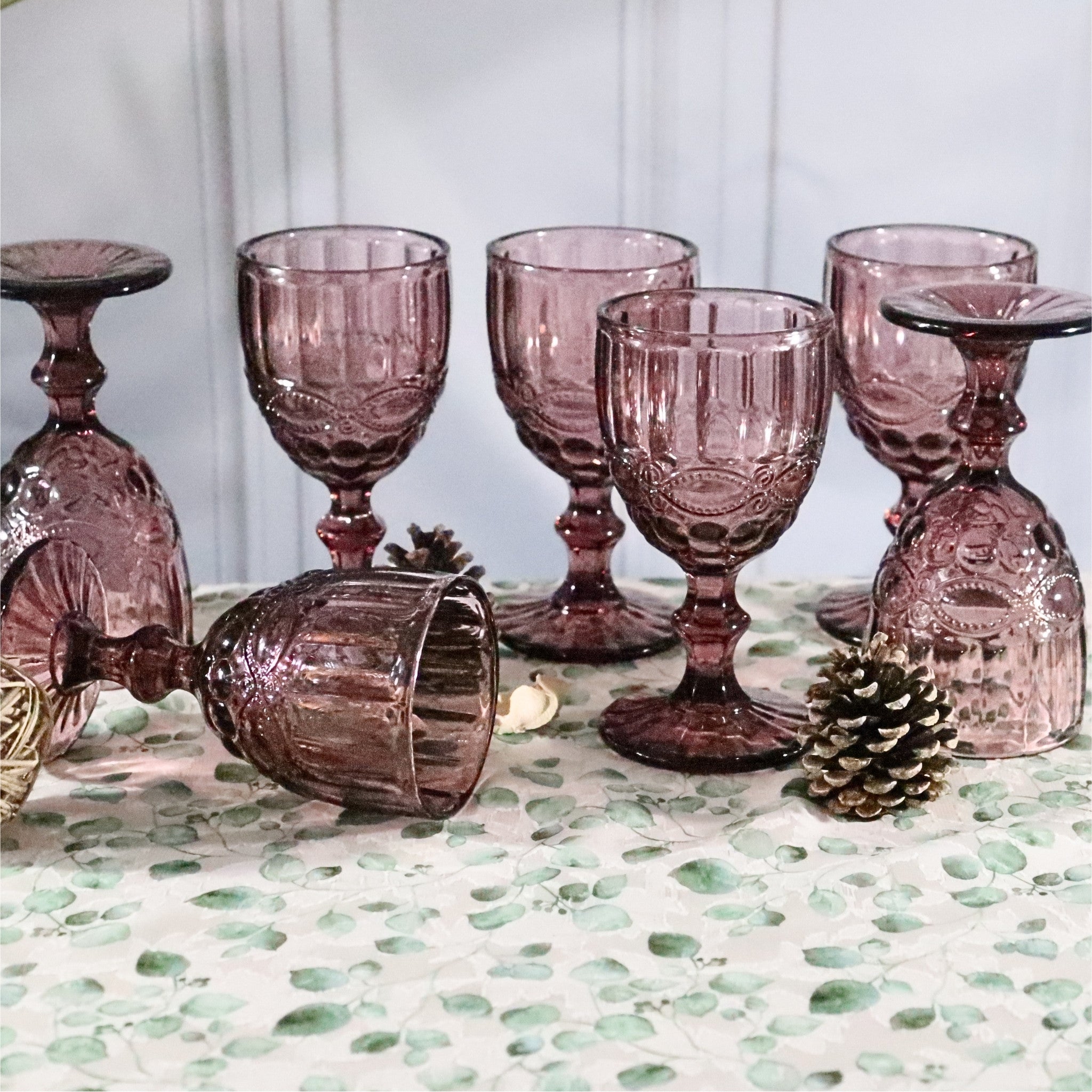 Set of purple glass goblets on a decorative surface with pine cones.
