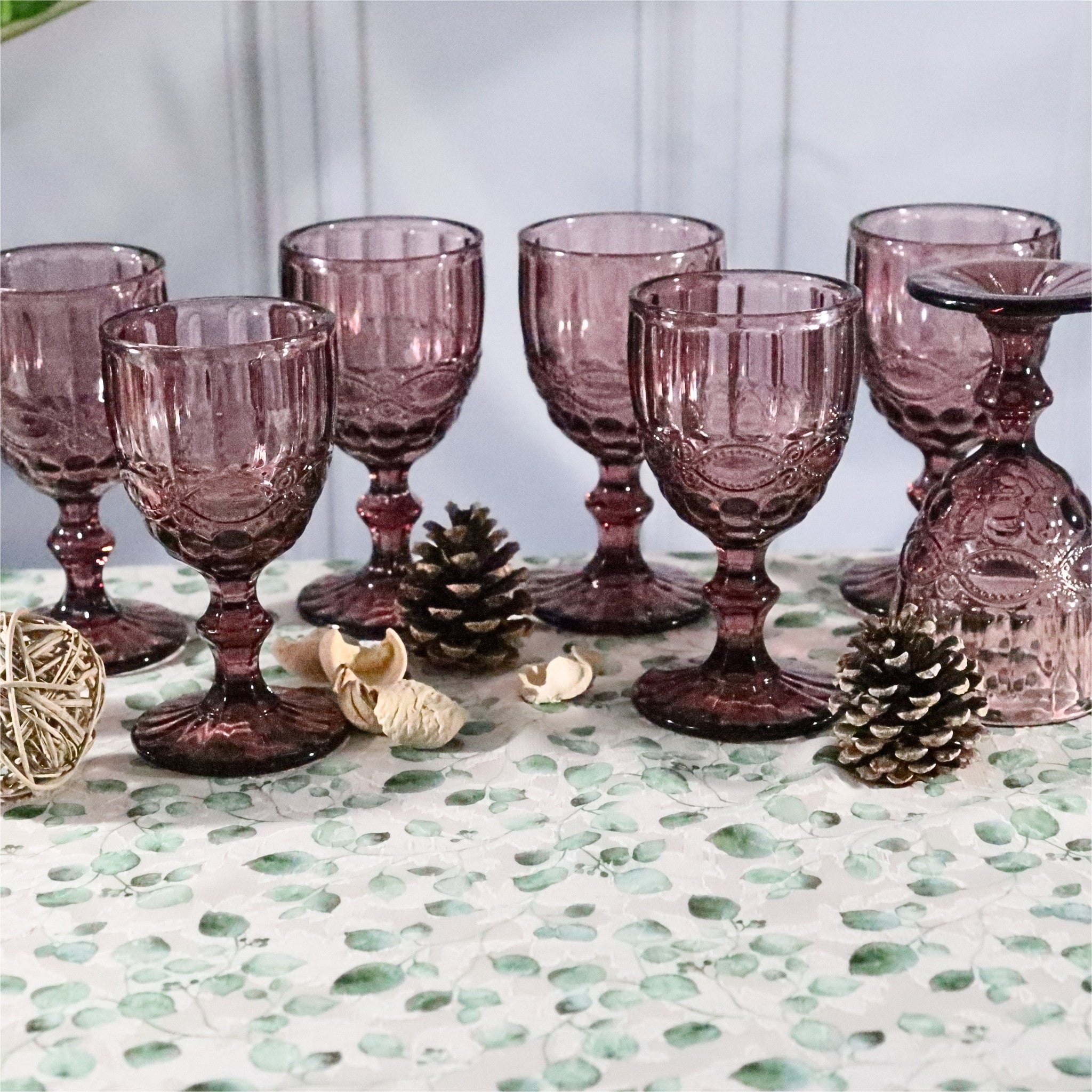 Set of purple glass goblets on a decorative table with pinecones and a textured surface.