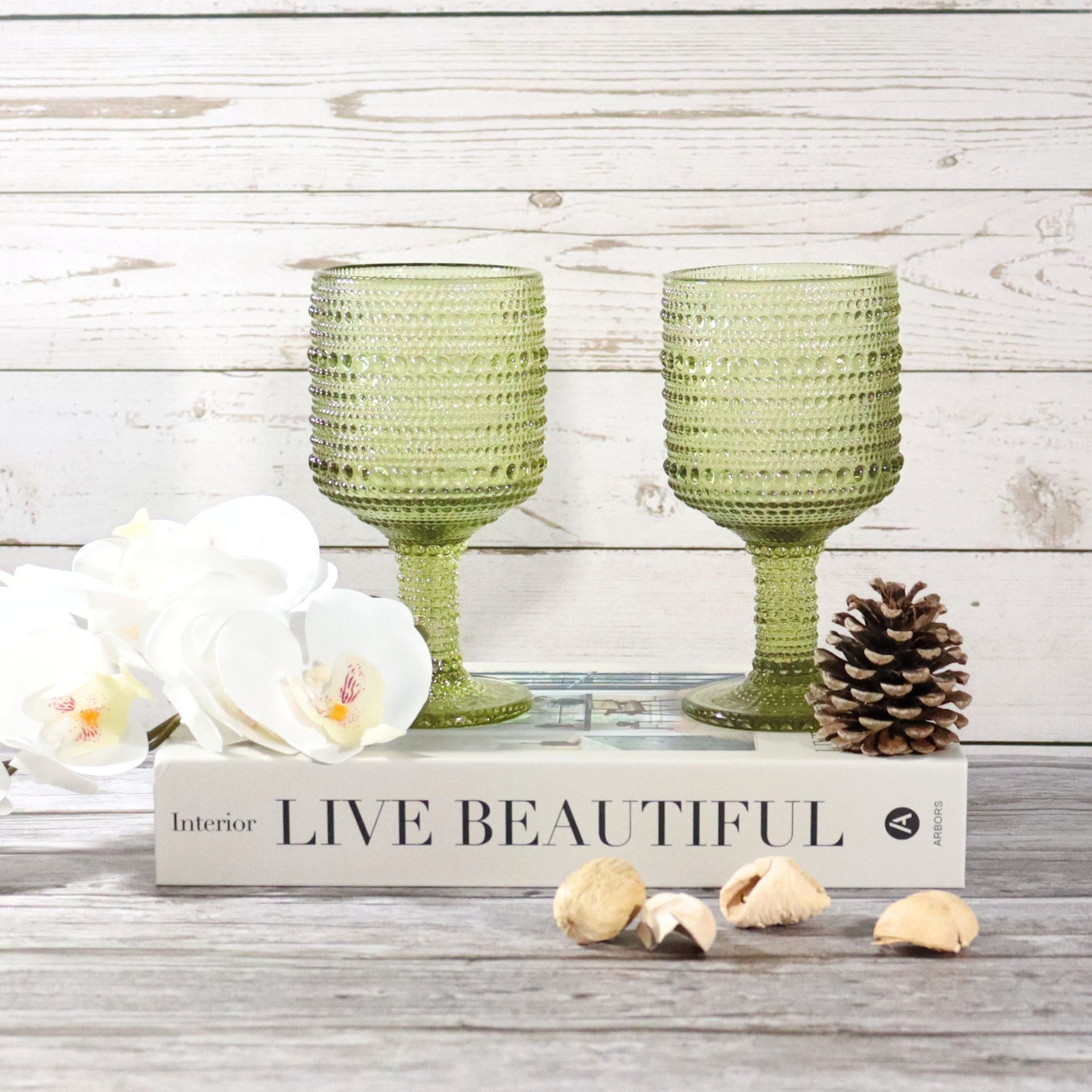 Two green textured glasses on a wooden block with 'Live Beautiful' text, surrounded by flowers and pinecones.