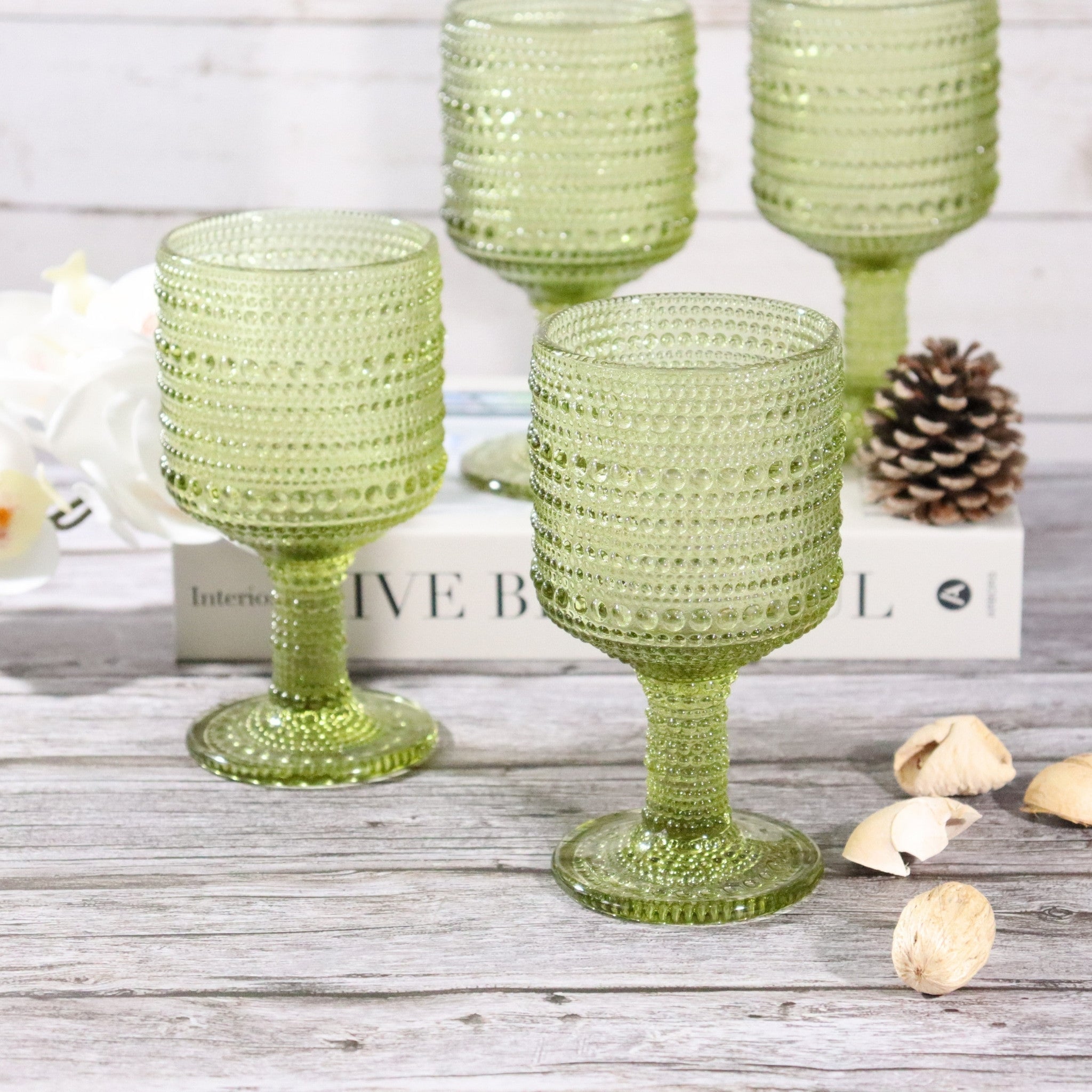 Set of green textured glass goblets on a wooden surface with pinecones and books in the background.