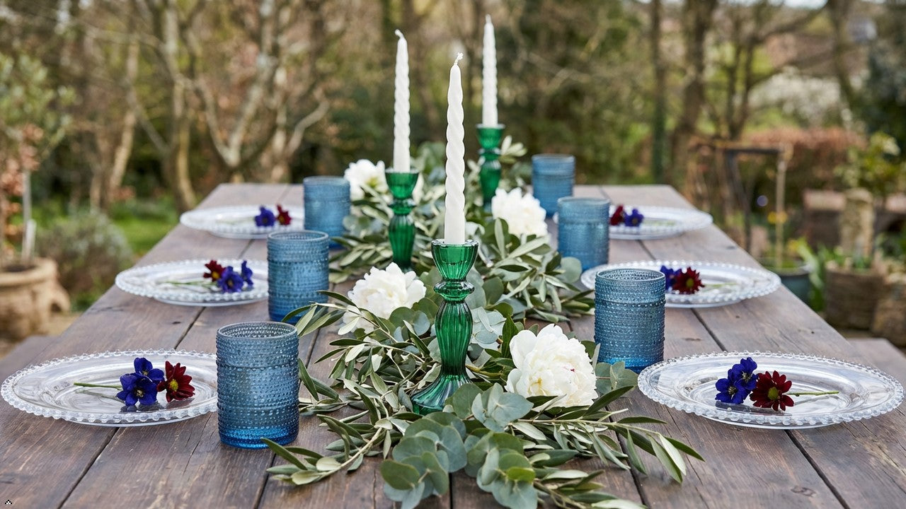 Decorative table setting with candles, blue glassware, and flowers on a wooden table outdoors.
