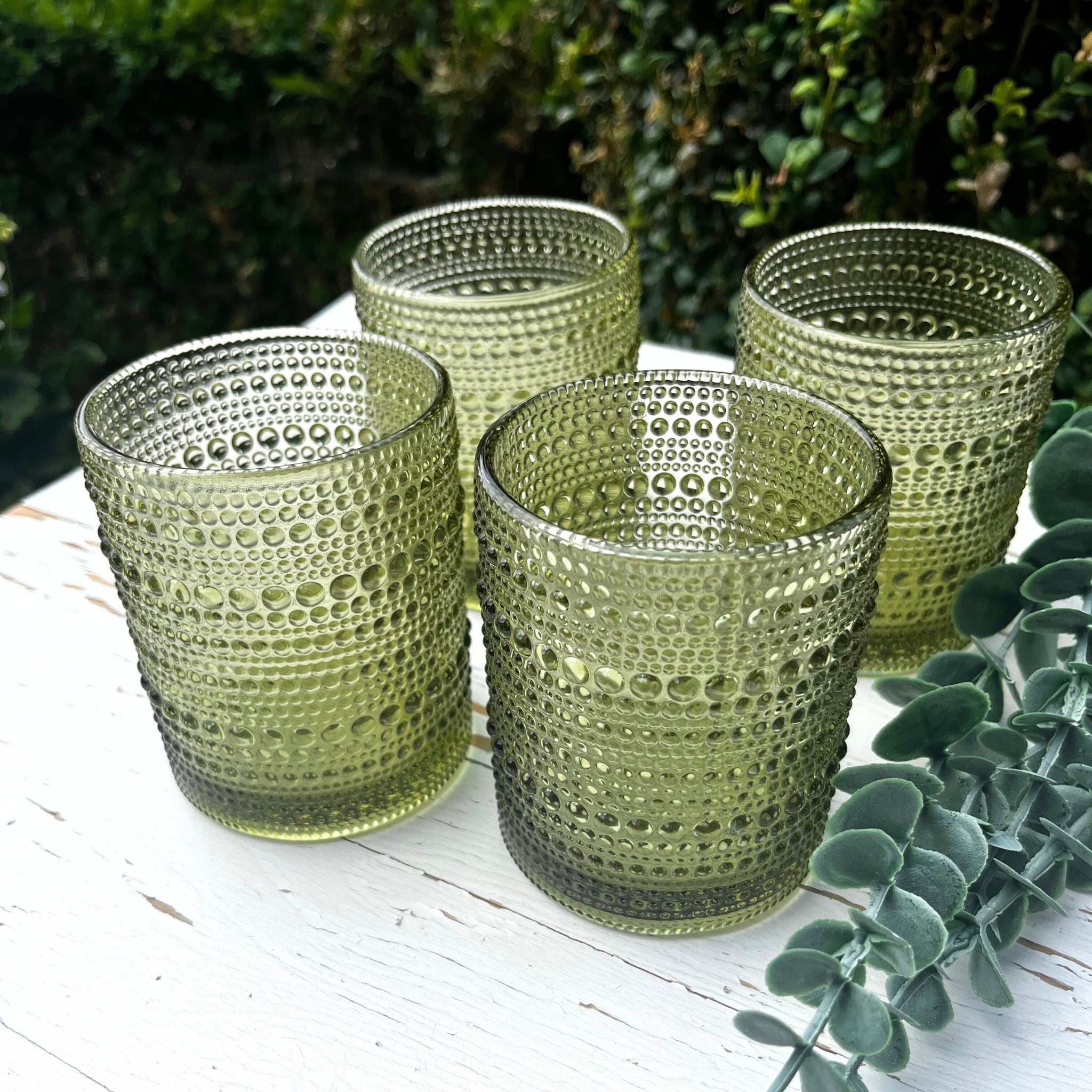 Four green hobnail glass tumblers on a white surface with greenery in the background