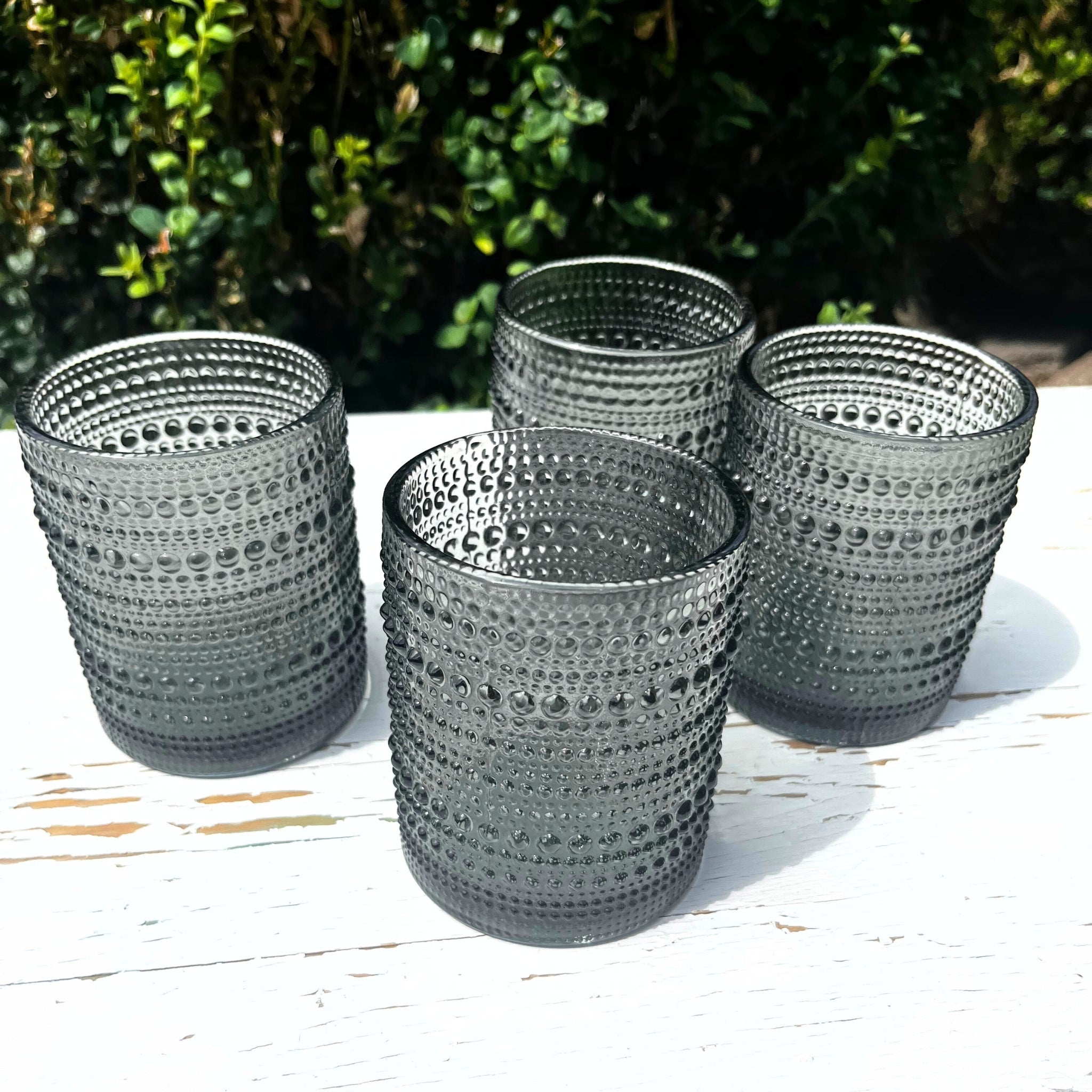 Four hobnail gray glass tumblers on a wooden surface with a blurred green background