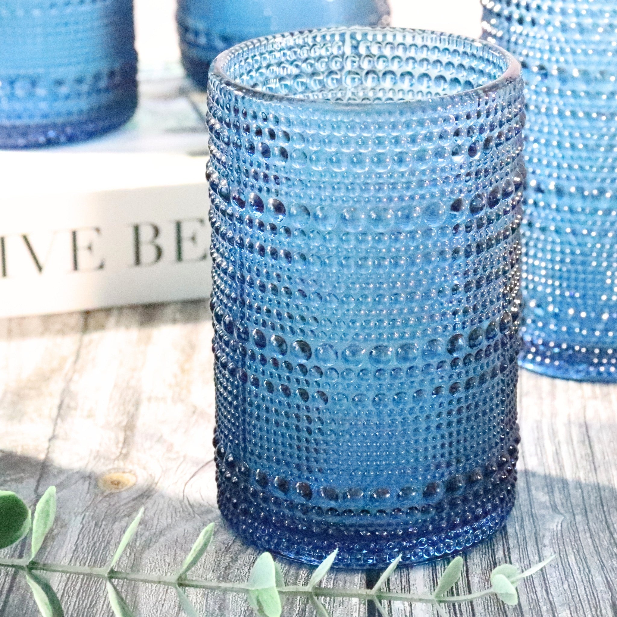 Blue hobnail jupiter bead drinking glasses on a wooden surface with other similar glasses in the background.
