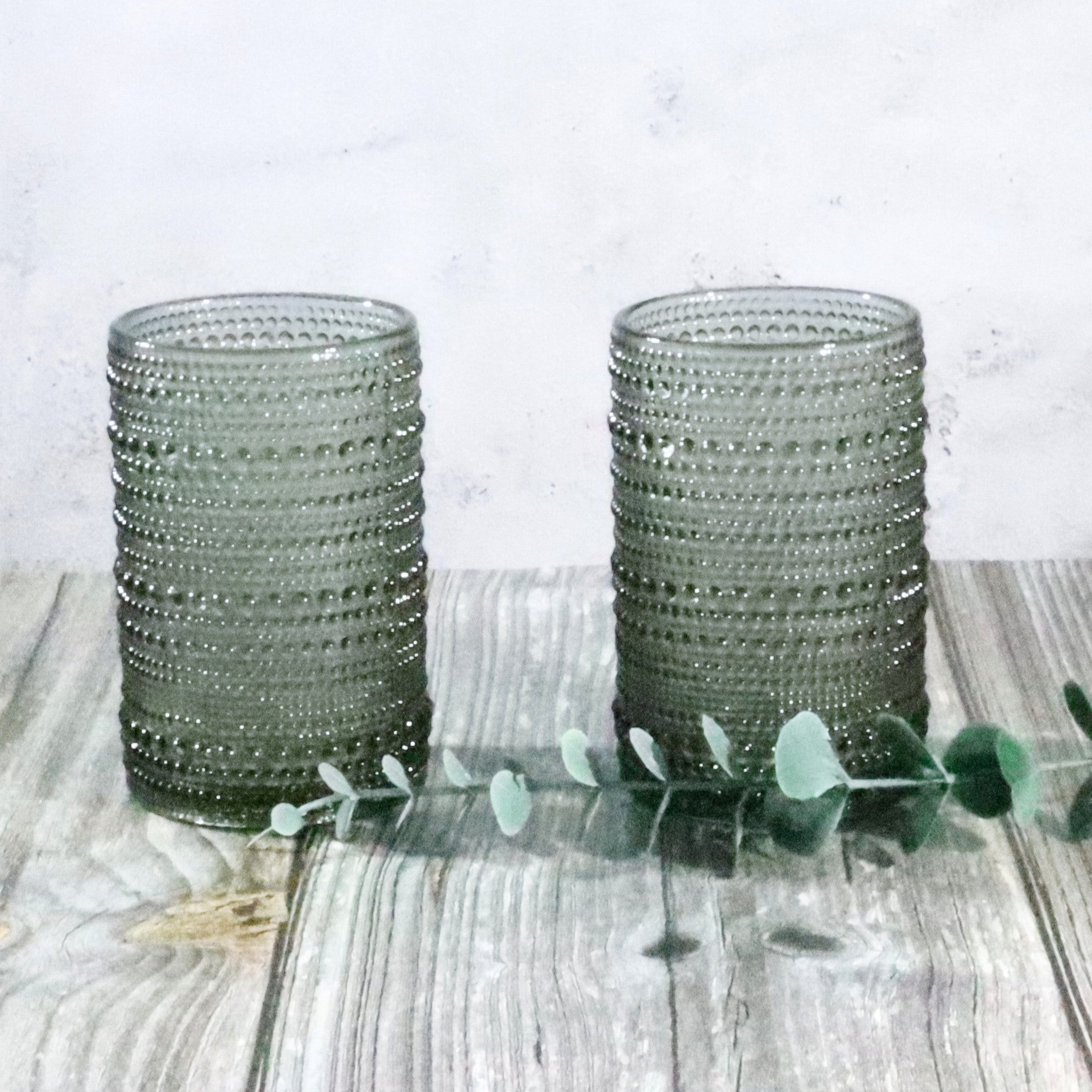 Two grey hobnail bead glass tumblers on a wooden surface with a white background