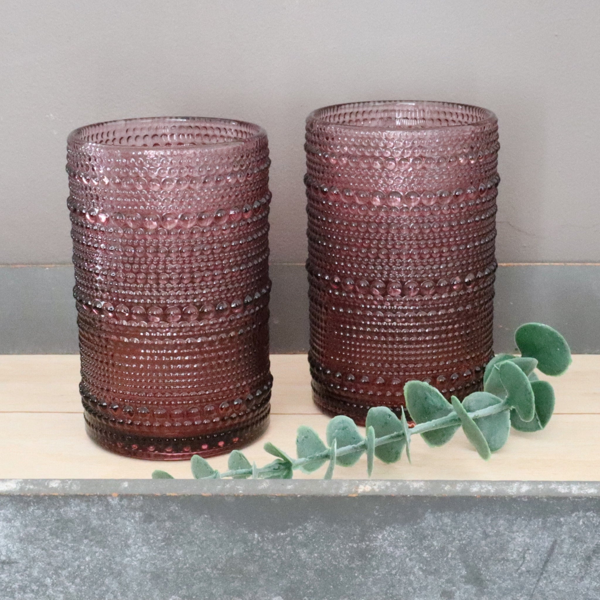 Two hobnail textured purple glass vases on a wooden surface with a plant leaf.