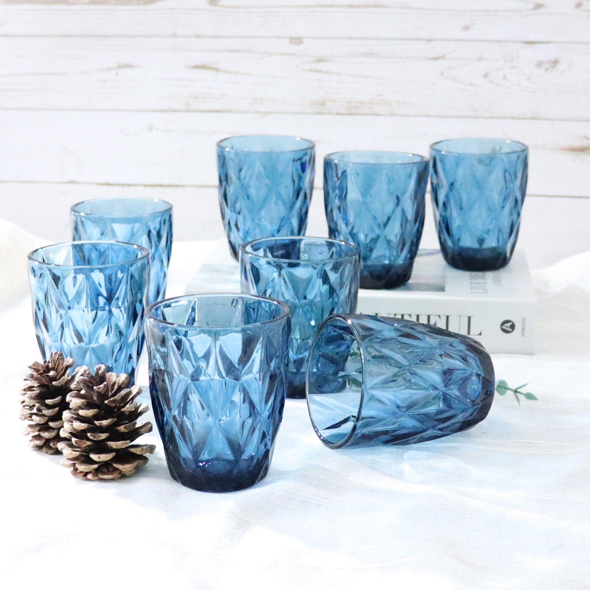 Set of blue glass tumblers on a white surface with a pine cone and book in the background.