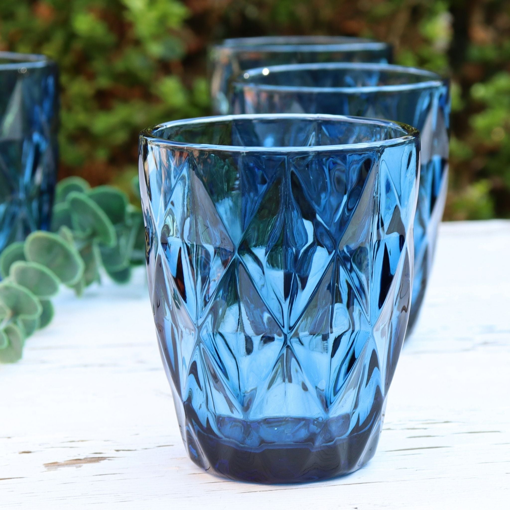 Set of blue glass tumblers with diamond pattern on a white surface with greenery in the background