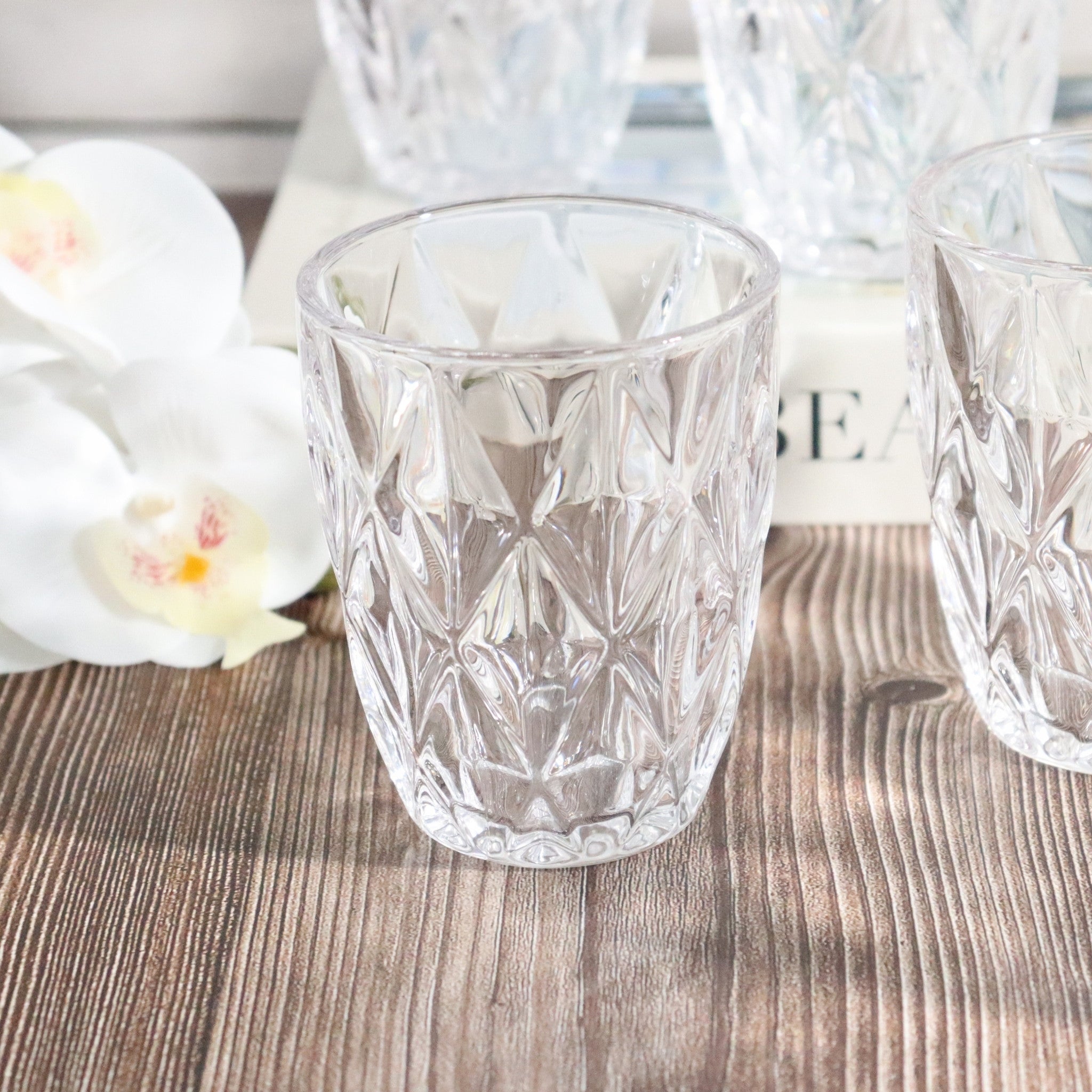 Clear glass tumblers with diamond cut pattern on a wooden surface with flowers and text in the background
