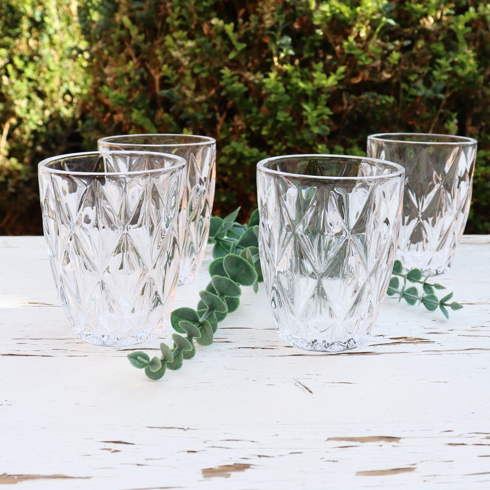 Four clear glass tumblers on a white surface with greenery in the background