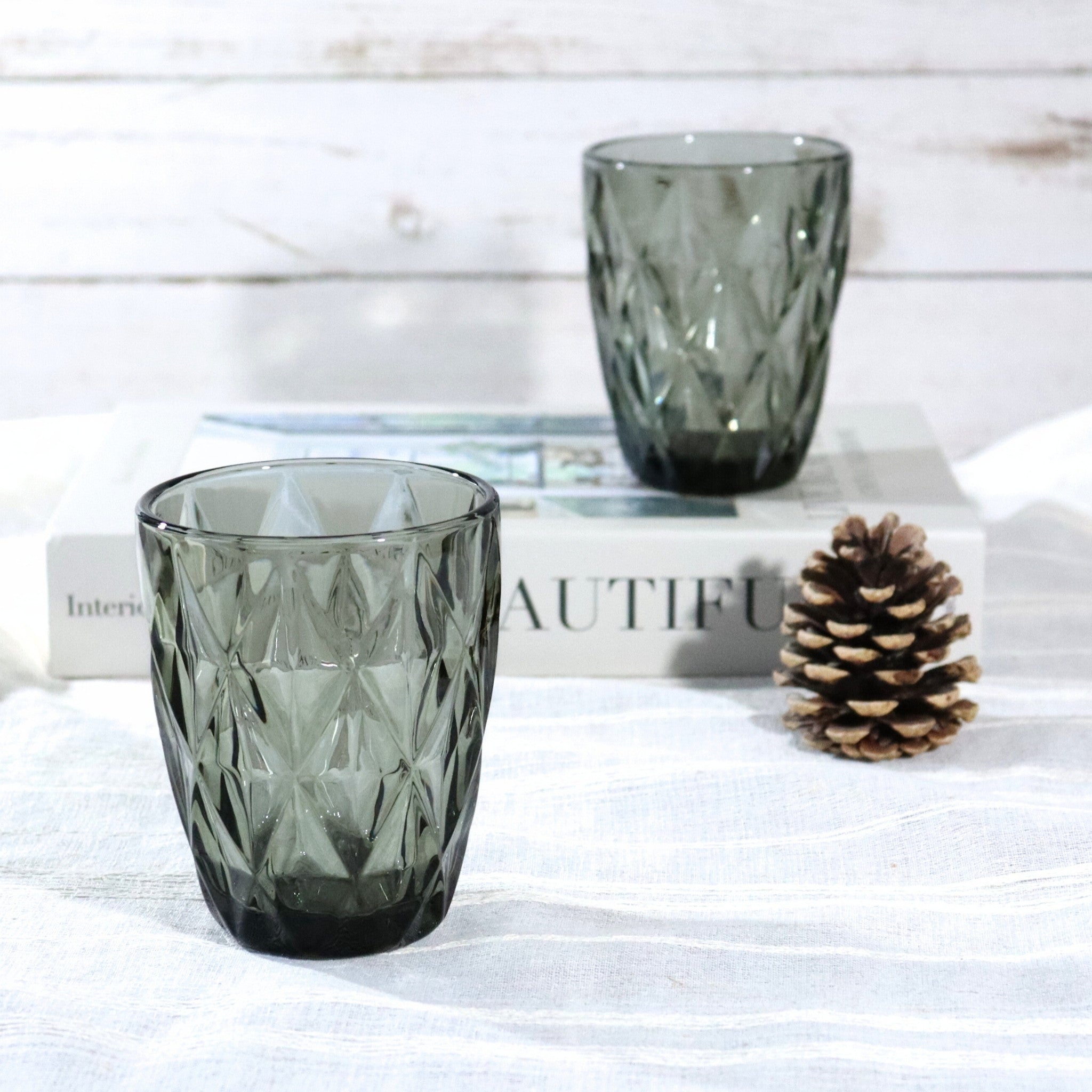 Two dark glass tumblers on books with a pine cone on a light wooden surface