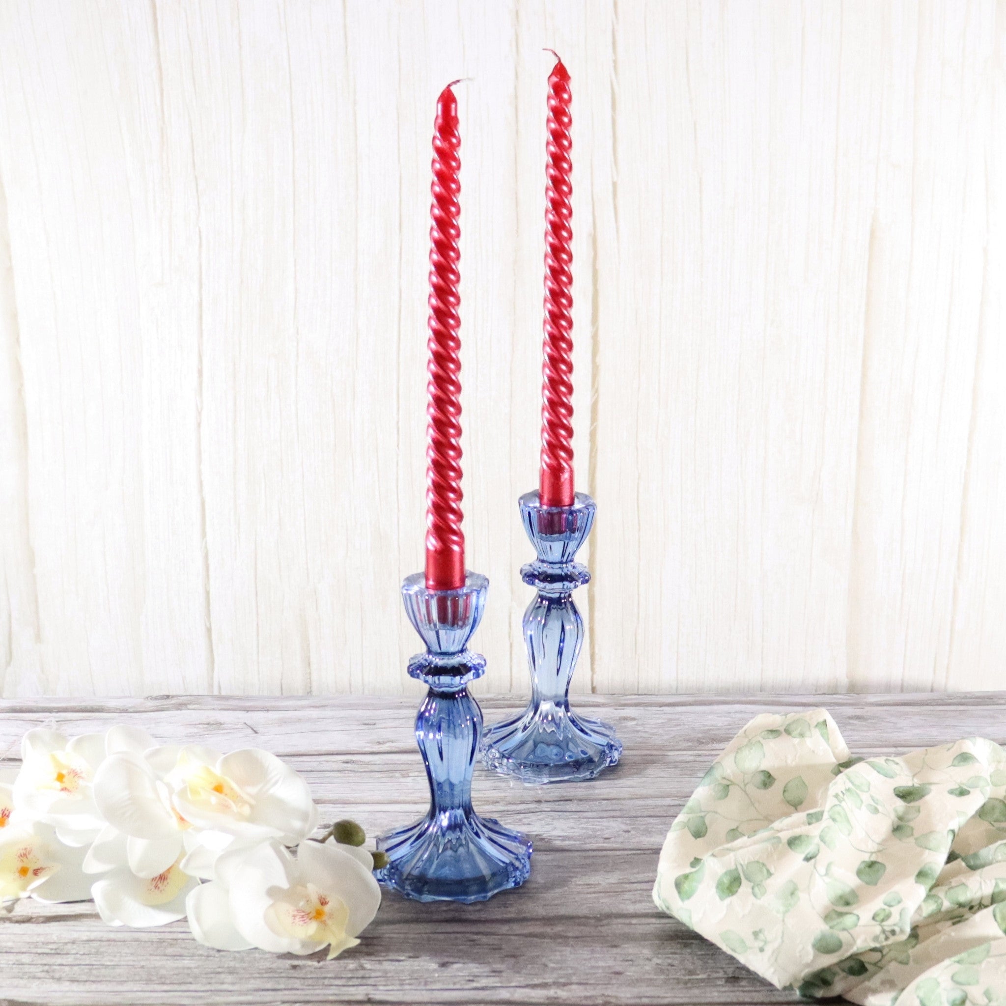 Two red spiral candles in a blue glass candleholder on a wooden surface with white flowers and greenery.