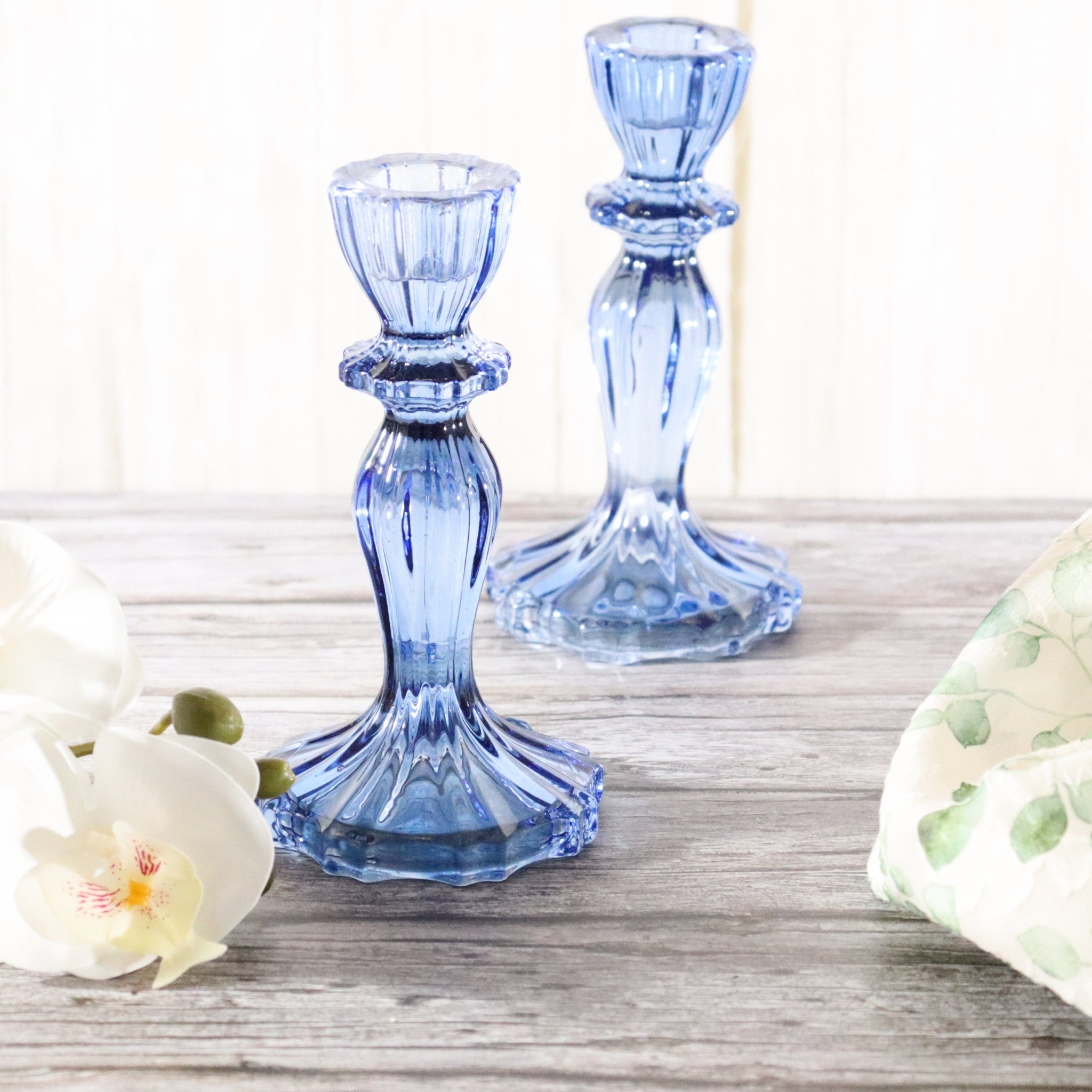 Two blue glass candle holders on a wooden surface with a white flower and greenery.