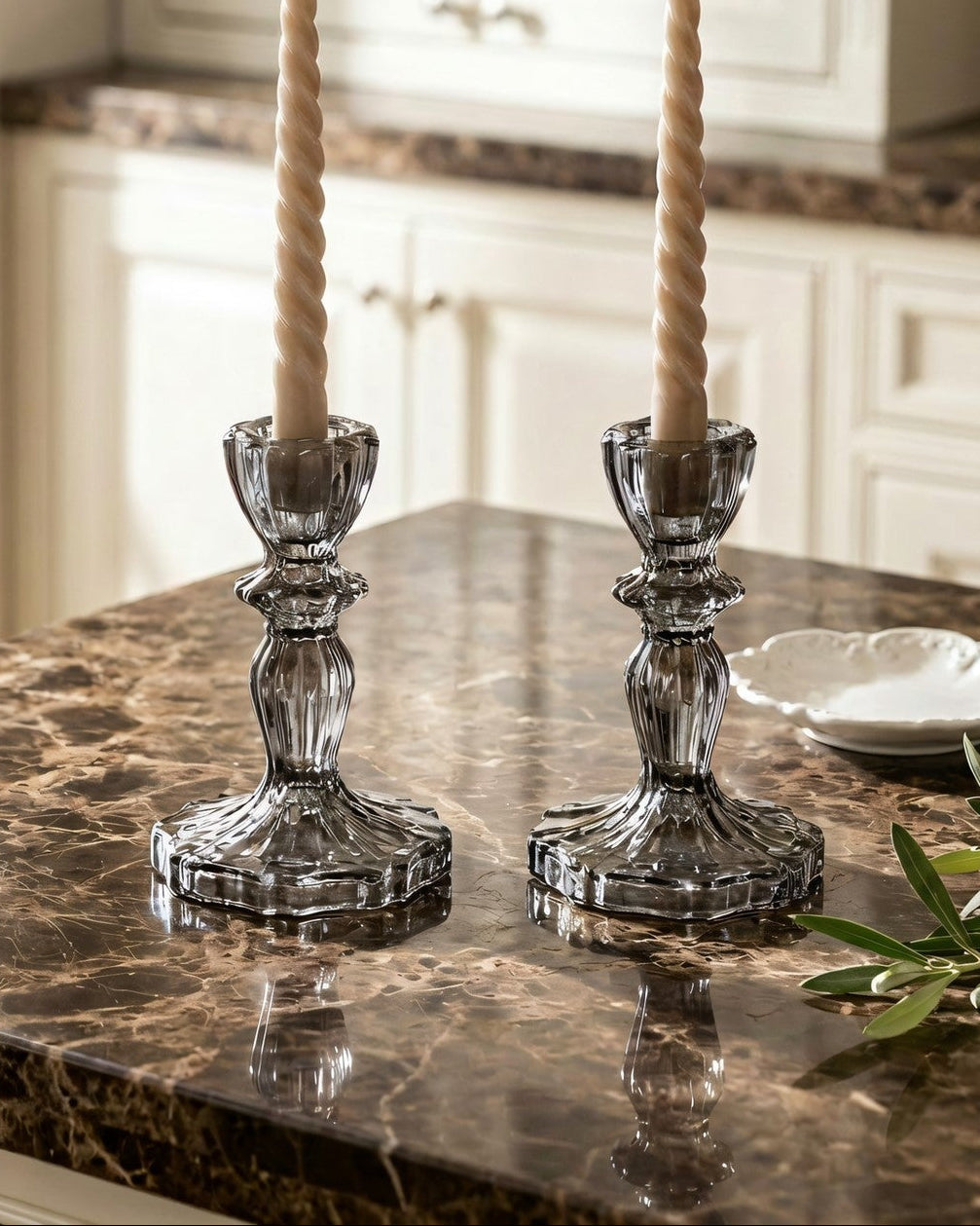 Two glass candlesticks with beige candles on a marble countertop in a kitchen.