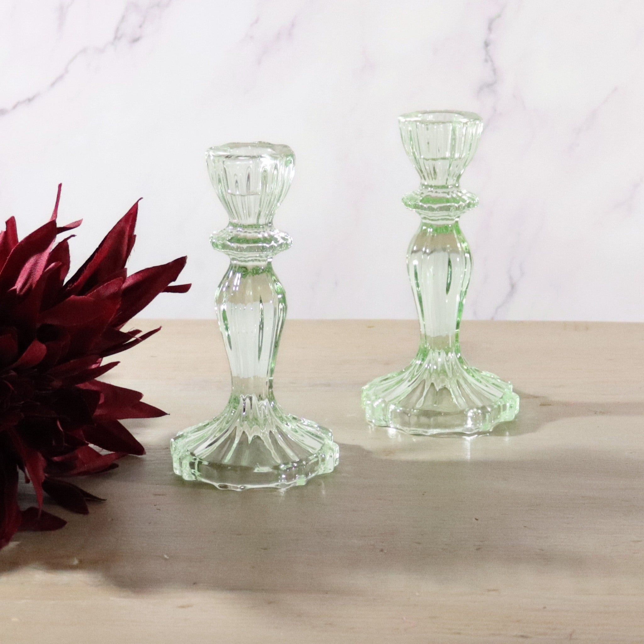 Two green clear glass candleholders on a wooden surface with a red flower in the background.