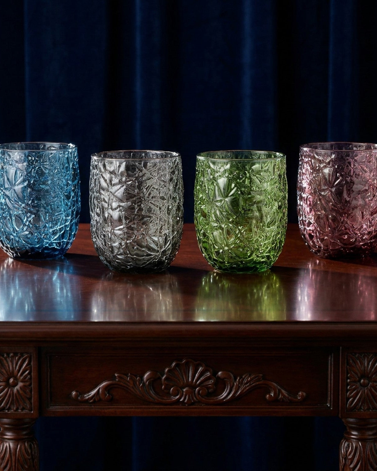 Four textured glass tumblers in blue, silver, green, and pink on a wooden table with a dark background.