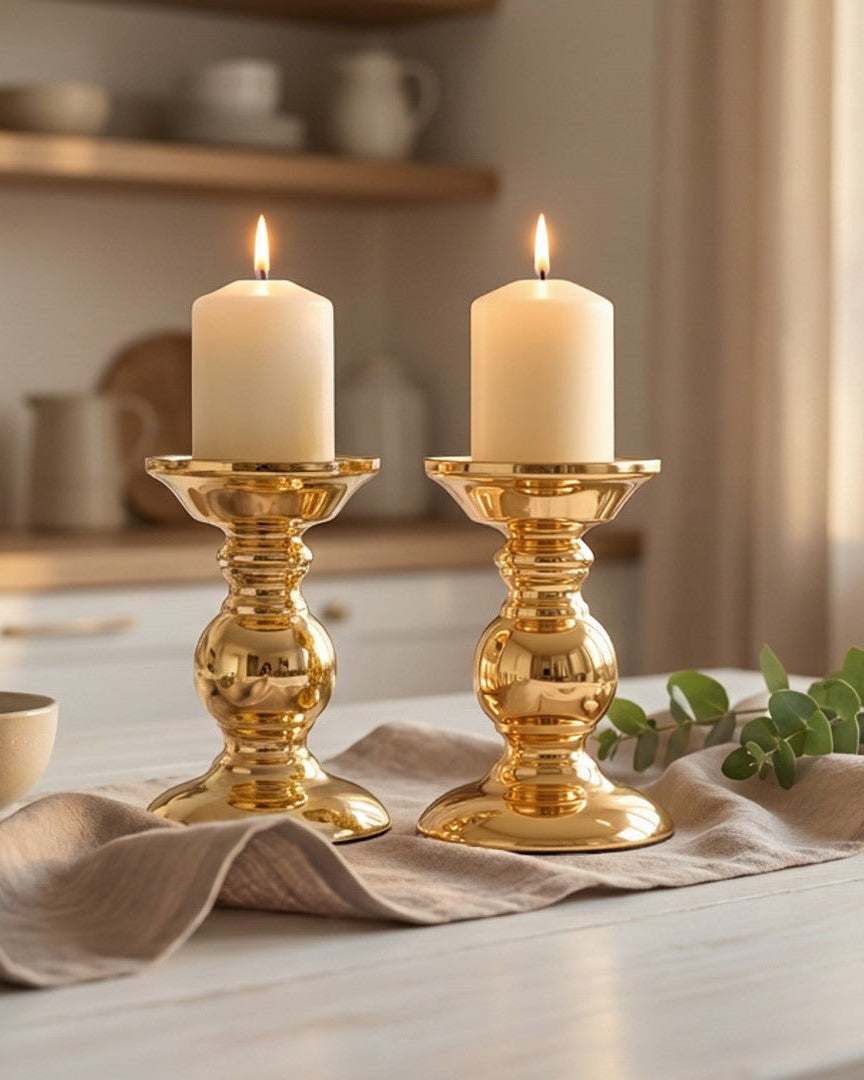 Two white candles in gold candle holders on a table with a kitchen background.