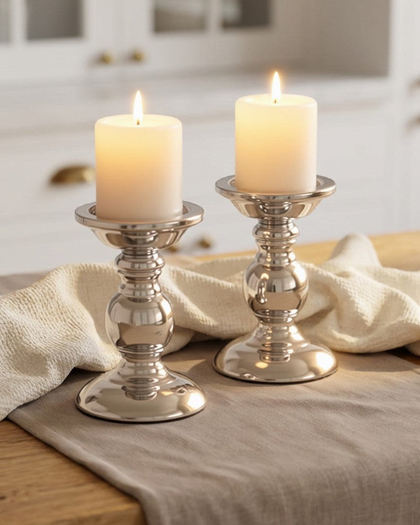Two candles in silver candlesticks on a wooden table with a kitchen background