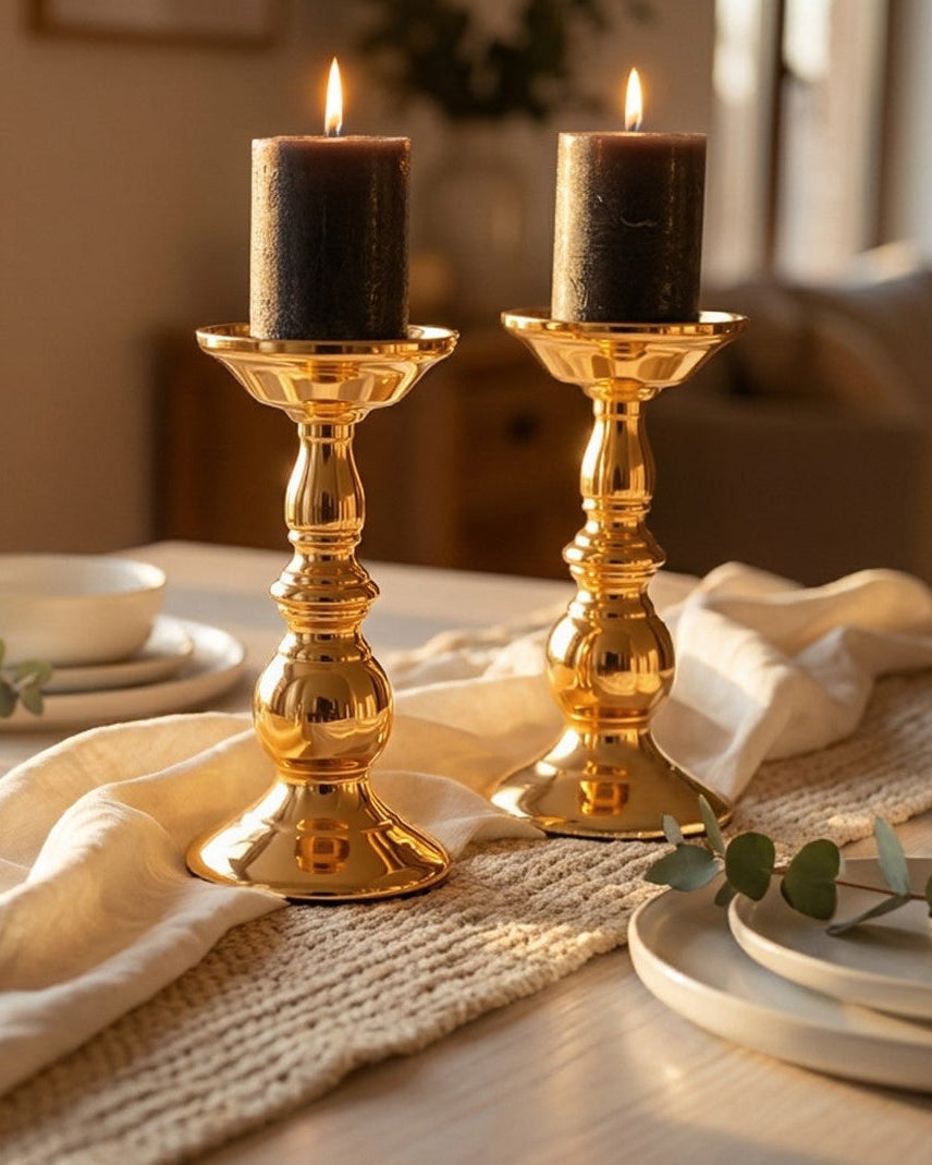 Two gold candlesticks with black candles on a table with a beige runner and white plates.