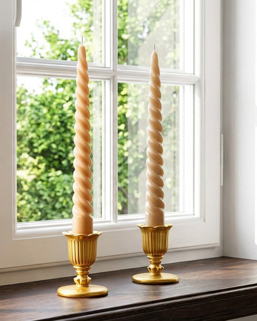 Two gold candle holders with spiral designs on a windowsill with greenery outside.