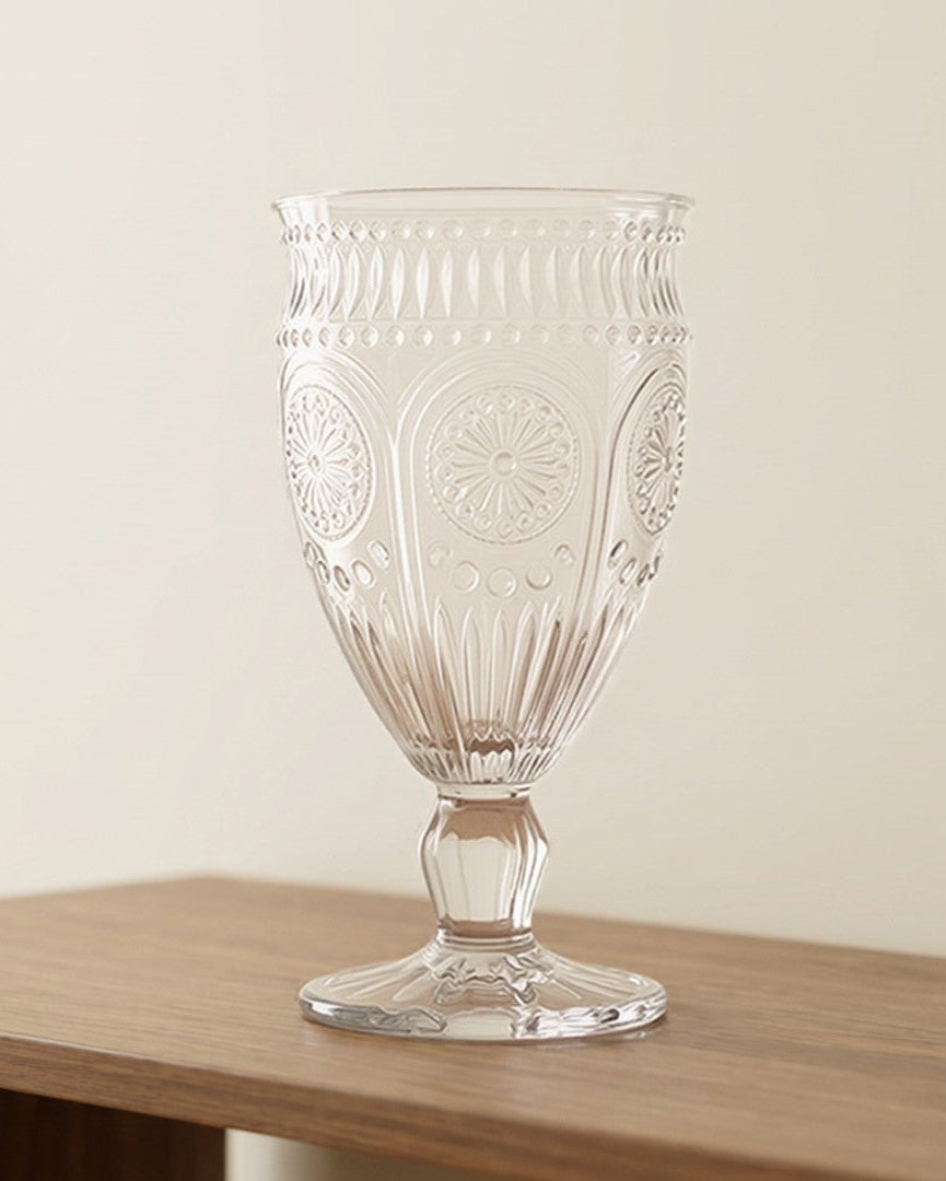 A clear glass goblet with etched floral patterns sits on a light brown wooden shelf against a warm beige wall.