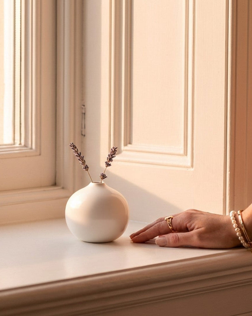 Person's hand near a small white vase with a plant on a windowsill.