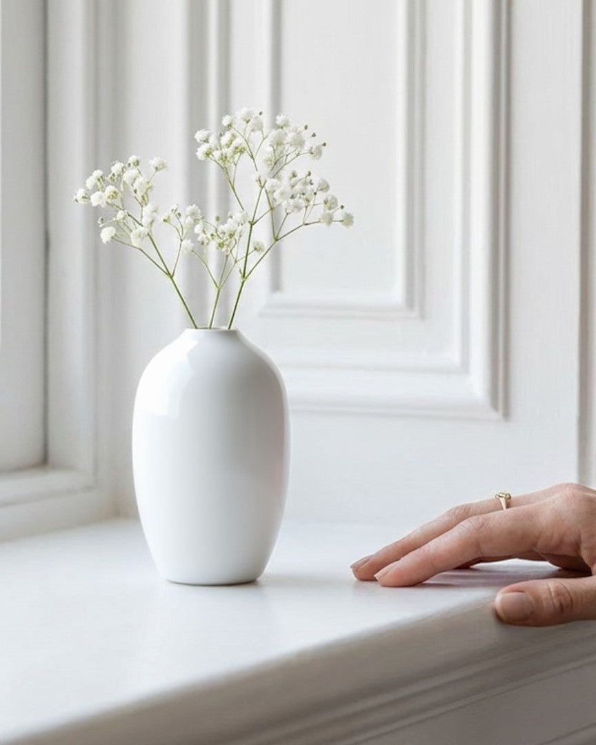 White vase with small white flowers on a windowsill with a hand reaching out.