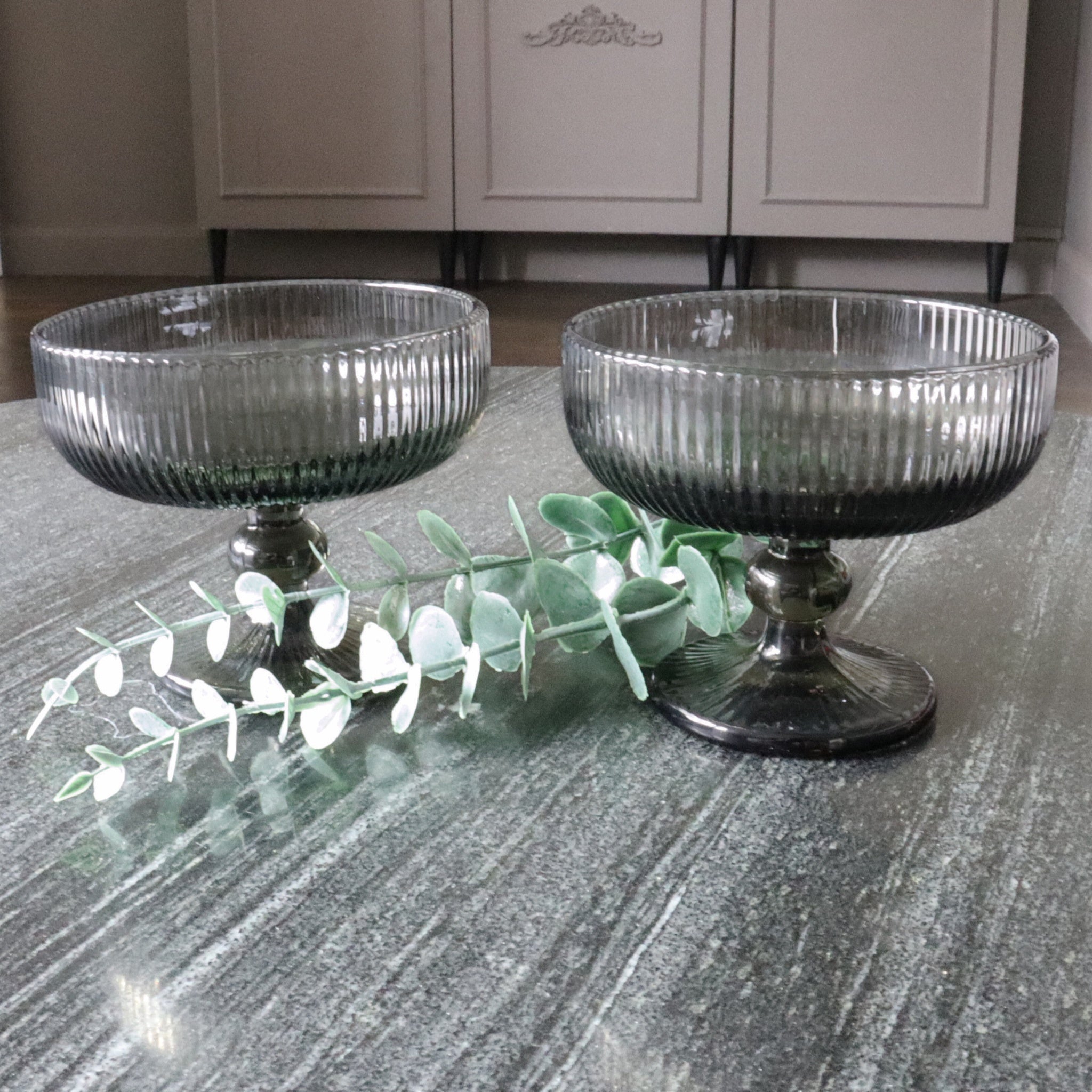 Two ribbed glass bowls on a textured surface with a branch of eucalyptus.