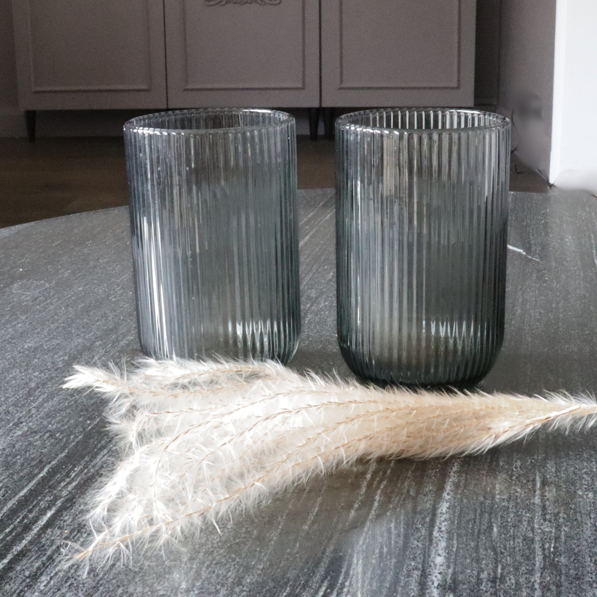 Two ribbed glass vases on a textured surface with decorative grasses.