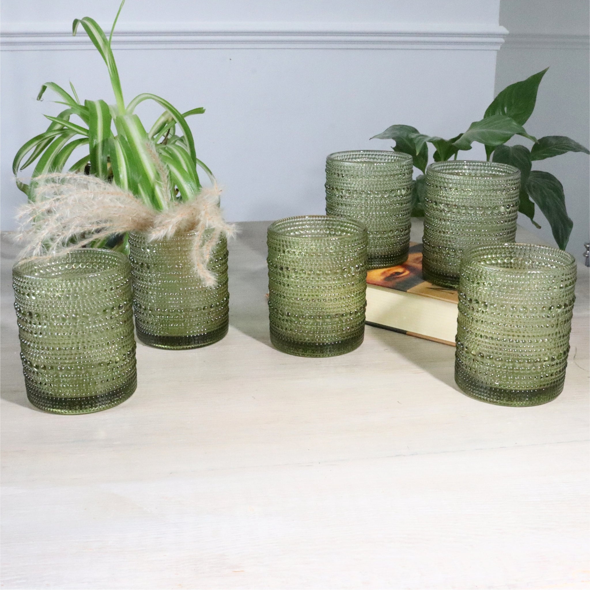 Set of green hobnail glass tumblers on a white surface with plants in the background
