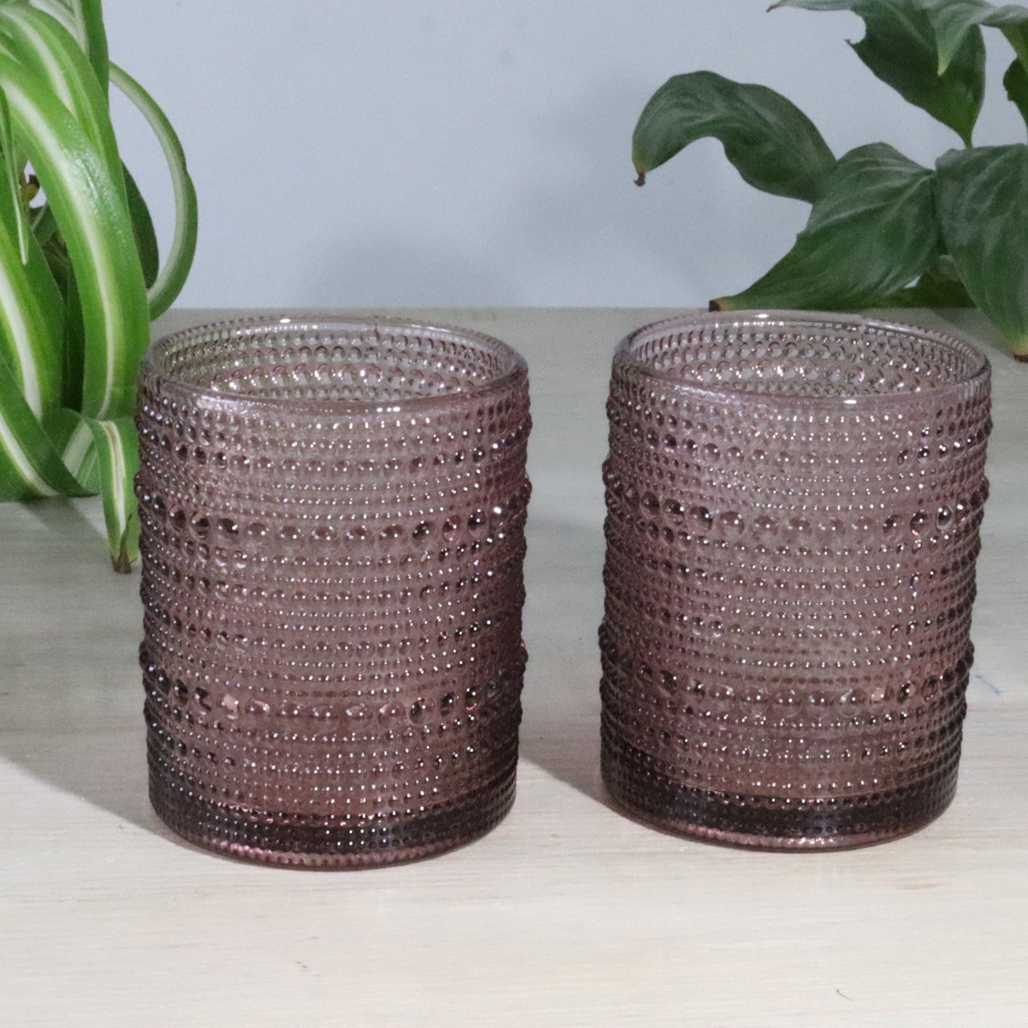 Two hobnail purple glass short tumblers on a light surface with plants in the background