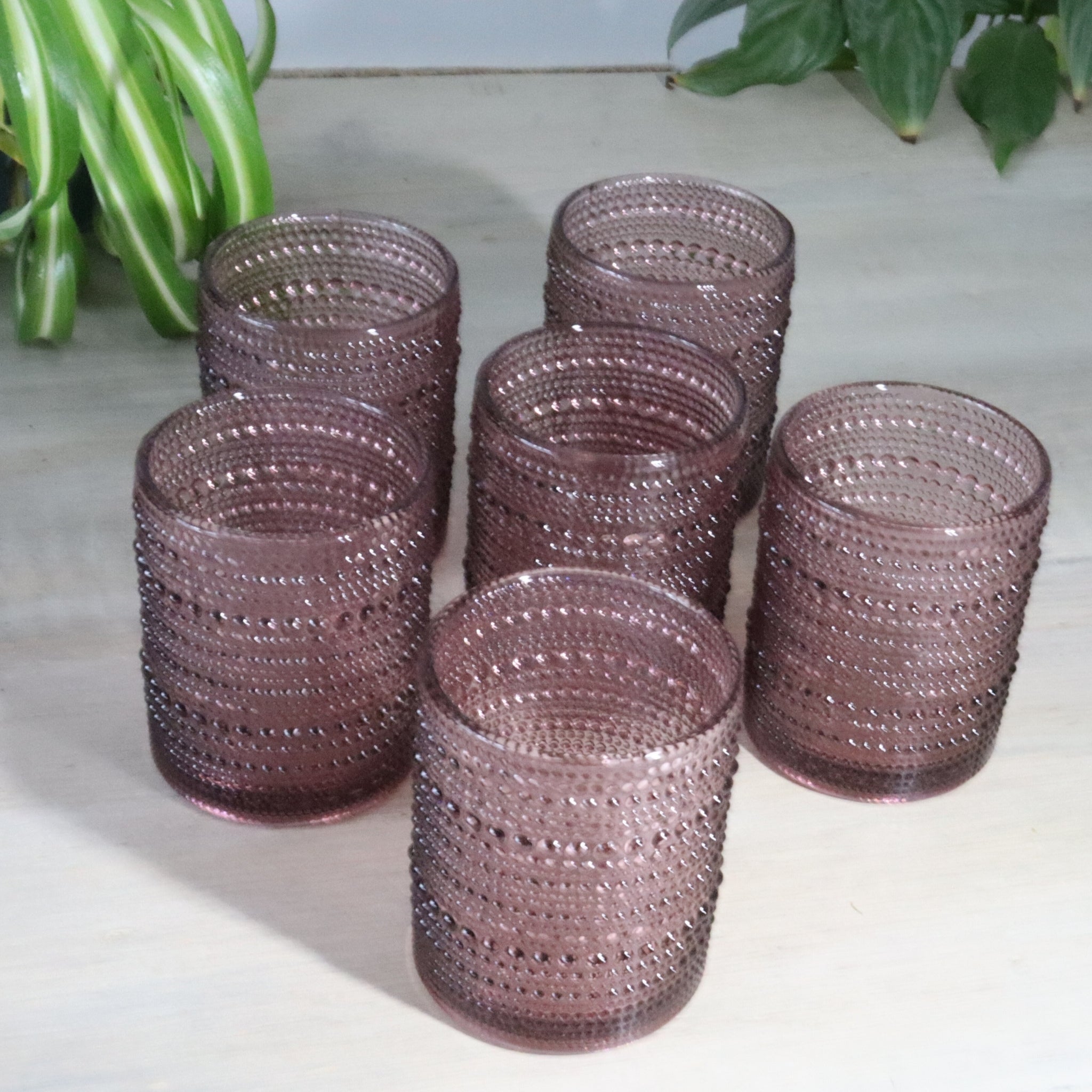 Set of purple hobnail glass tumblers on a light surface with green plants in the background