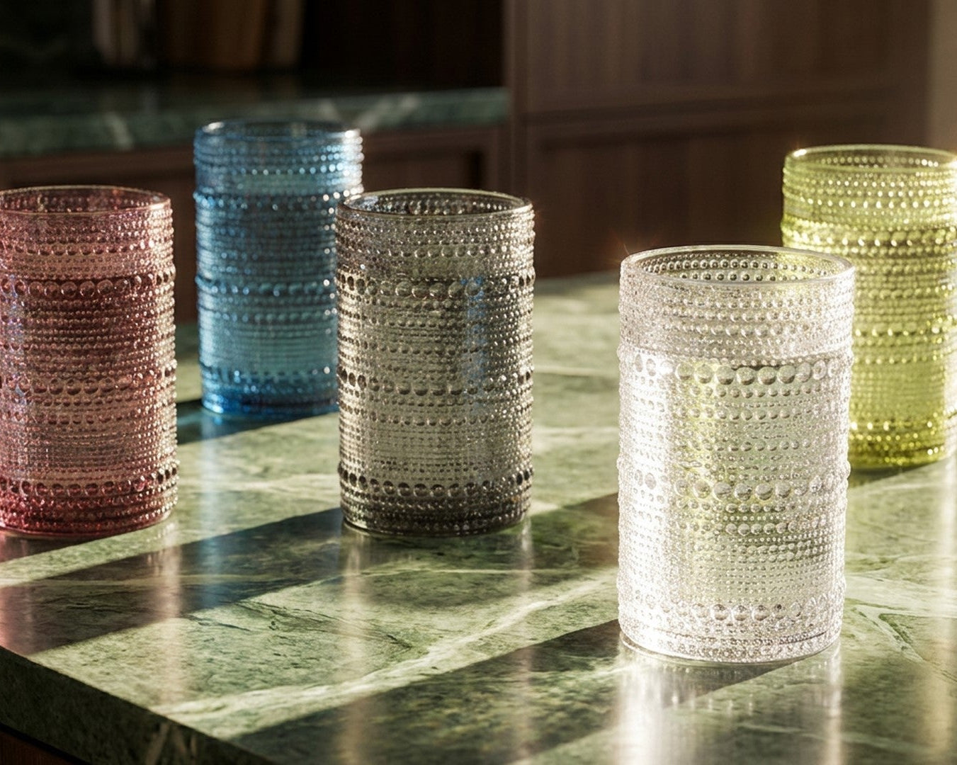 Set of five hobnail glass tumblers in various colours on a surface with plants in the background.