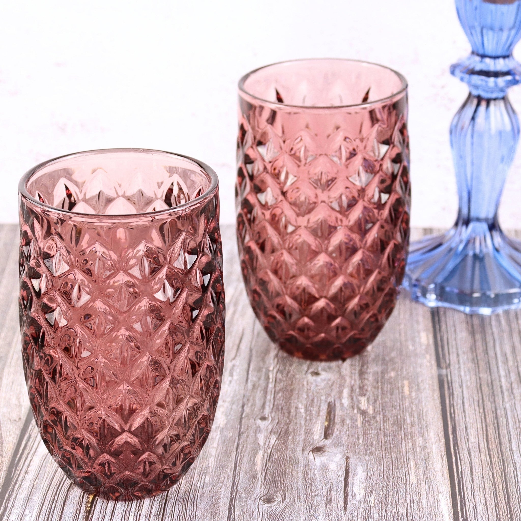 Two pink textured glass tumblers on a wooden surface with a blue glass object in the background.