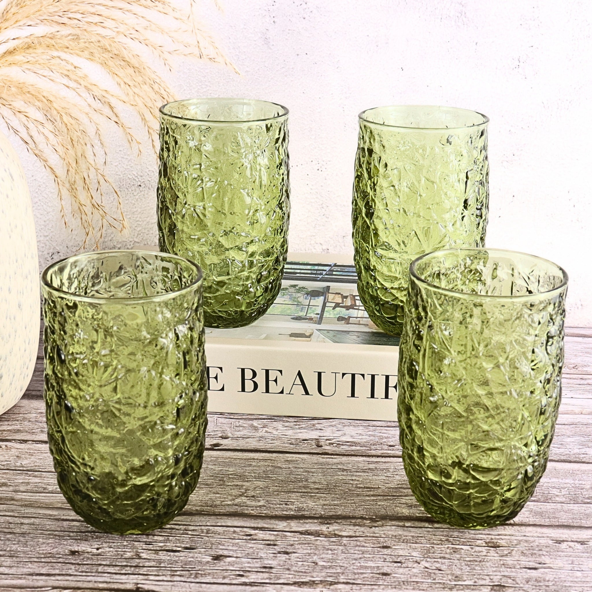 Four green textured glasses on a wooden surface with a decorative item in the background.